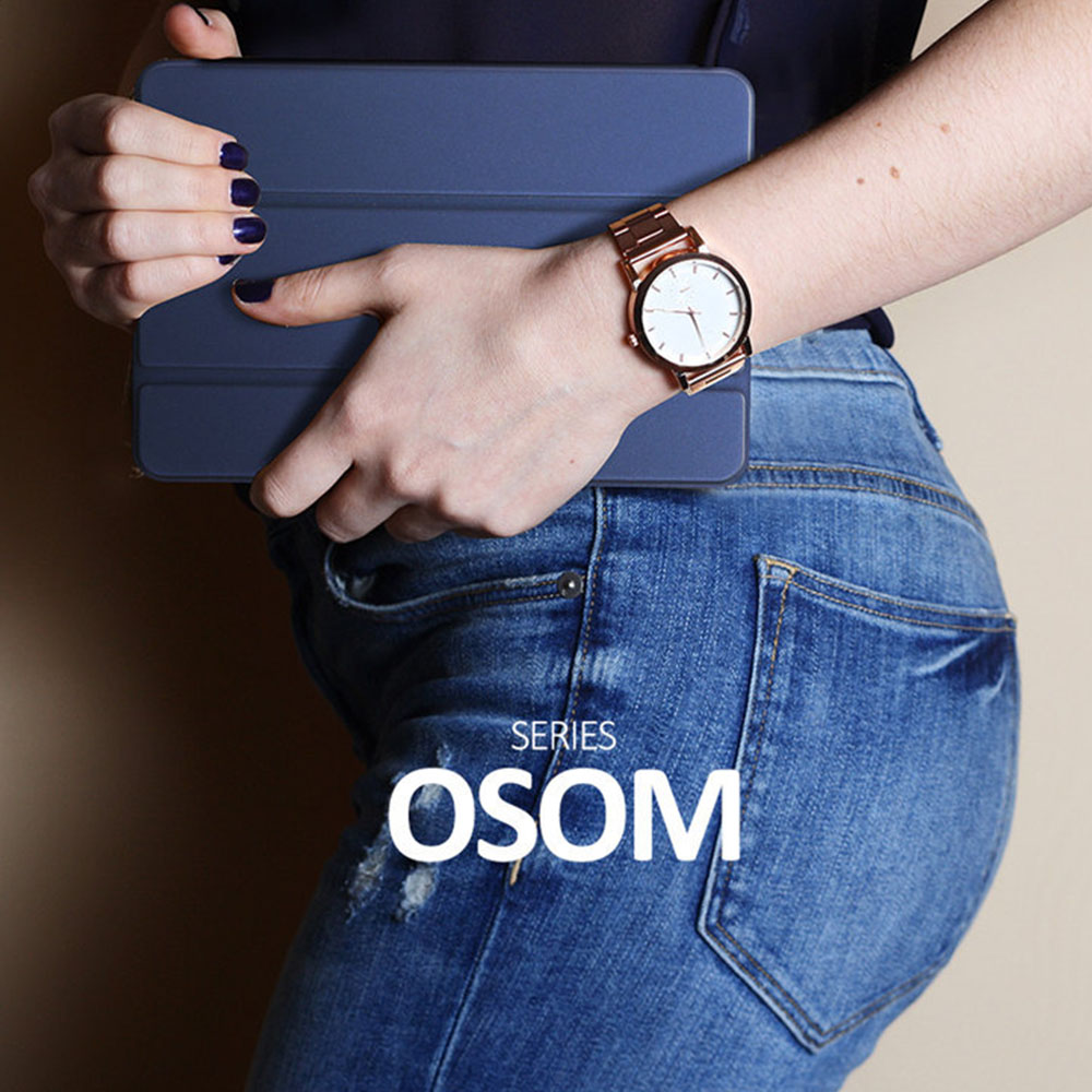 Eine Frau hält ein blaues Tablet mit einer goldenen Uhr am Handgelenk, trägt Jeans und ein dunkles Oberteil.