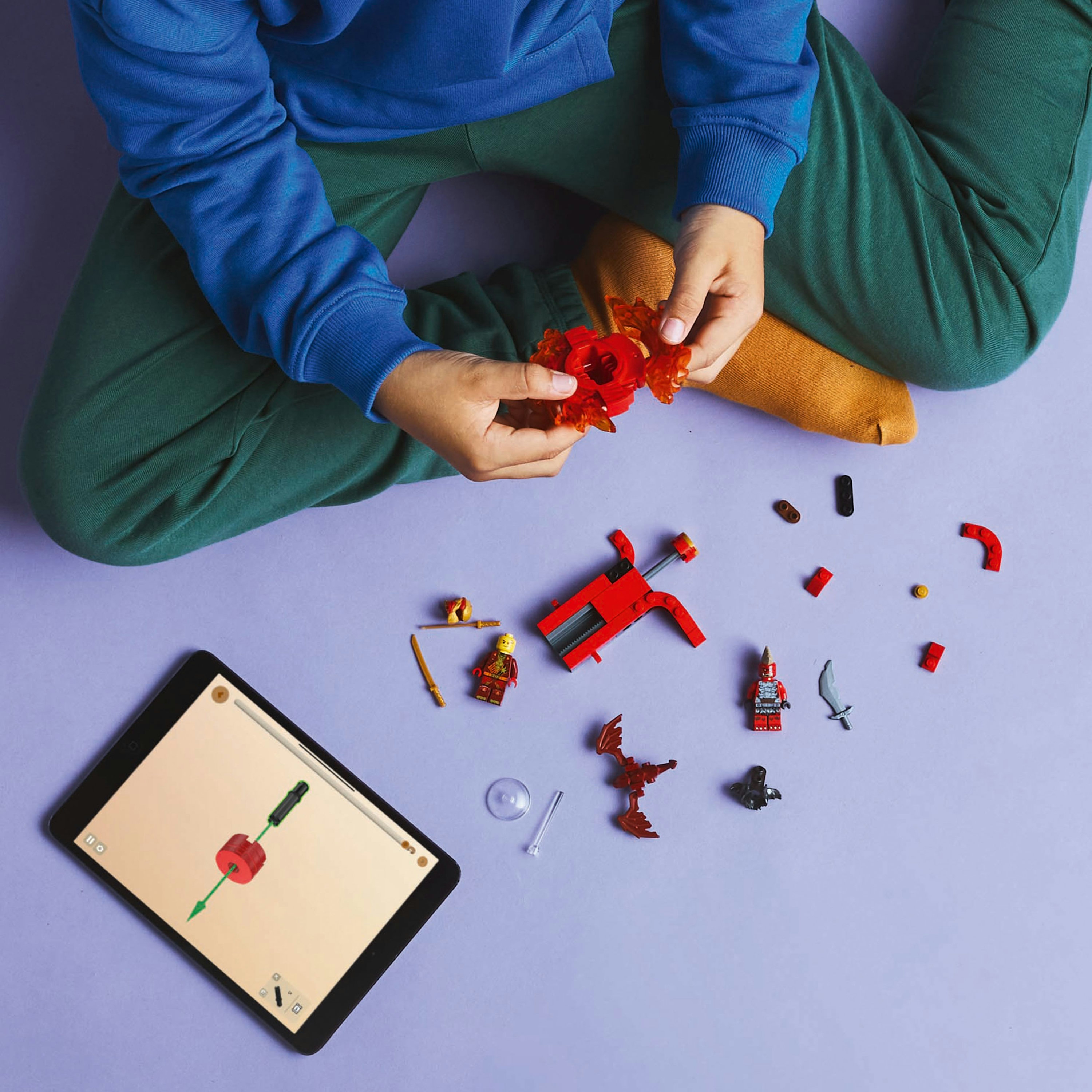 Bir kişi, kırmızı bir silah ve minifigürler dahil olmak üzere Lego parçalarını mor bir yüzeyde bir tablet ile birleştiriyor.