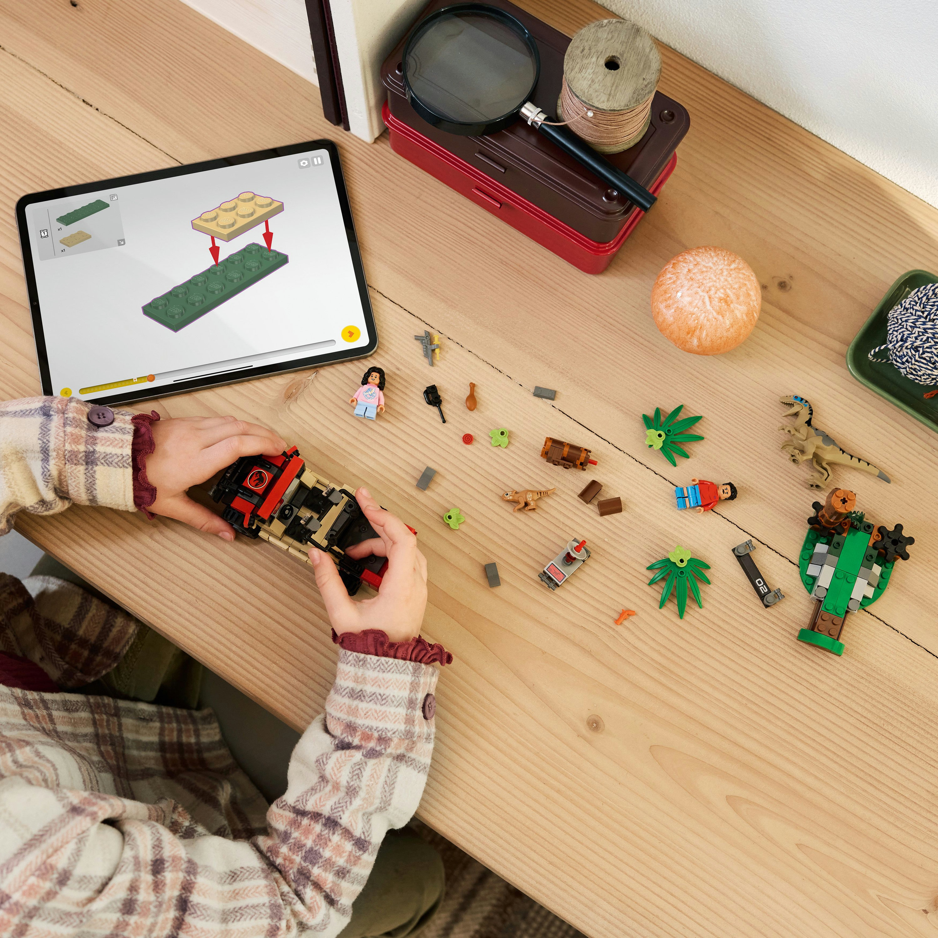 Zdjęcie z góry dziecka bawiącego się zestawem LEGO Jurassic World na drewnianym stole, korzystającego z instrukcji na tablecie.