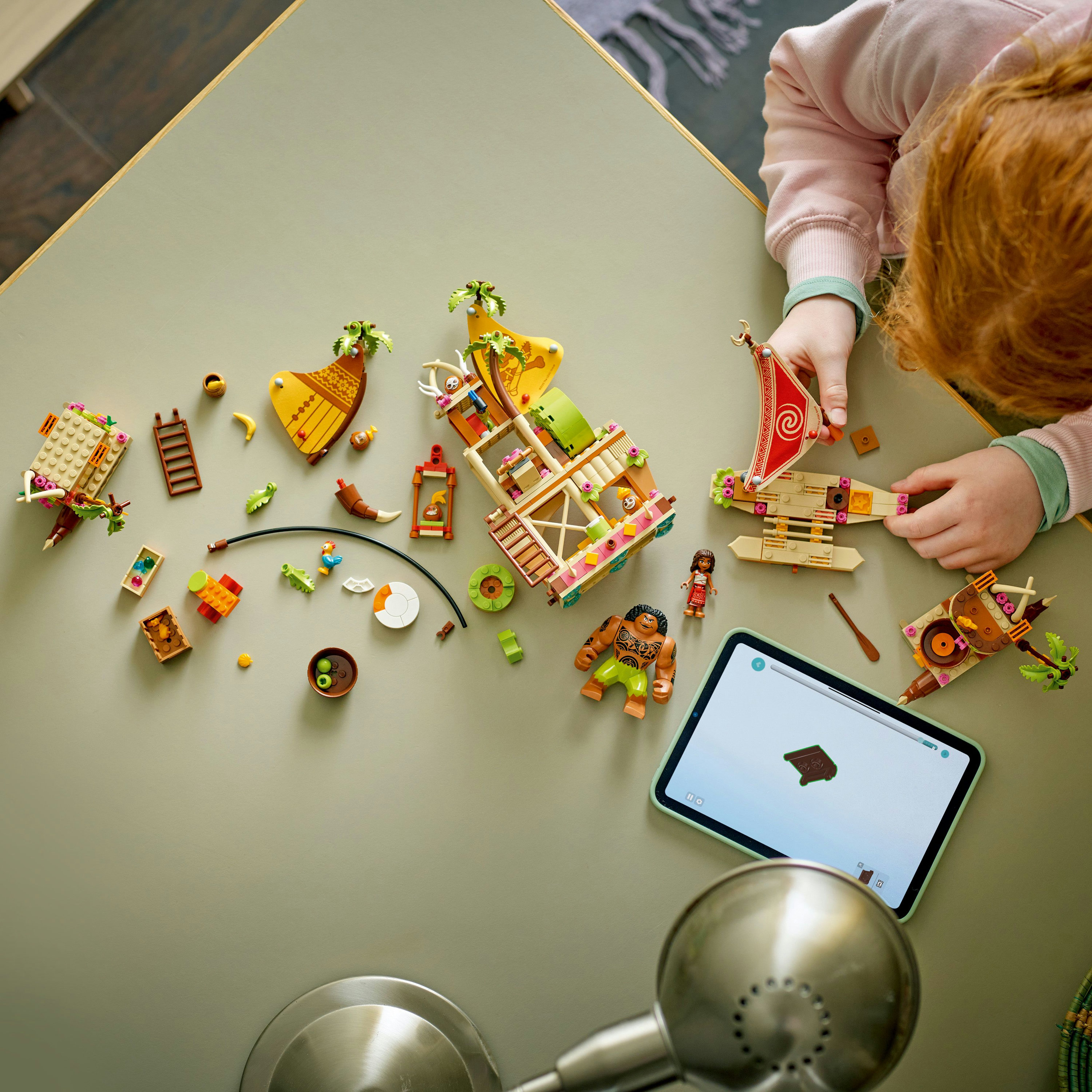 Widok z góry: Dziecko bawi się zestawem Lego Moana na zielonym stole, korzystając z tabletu.