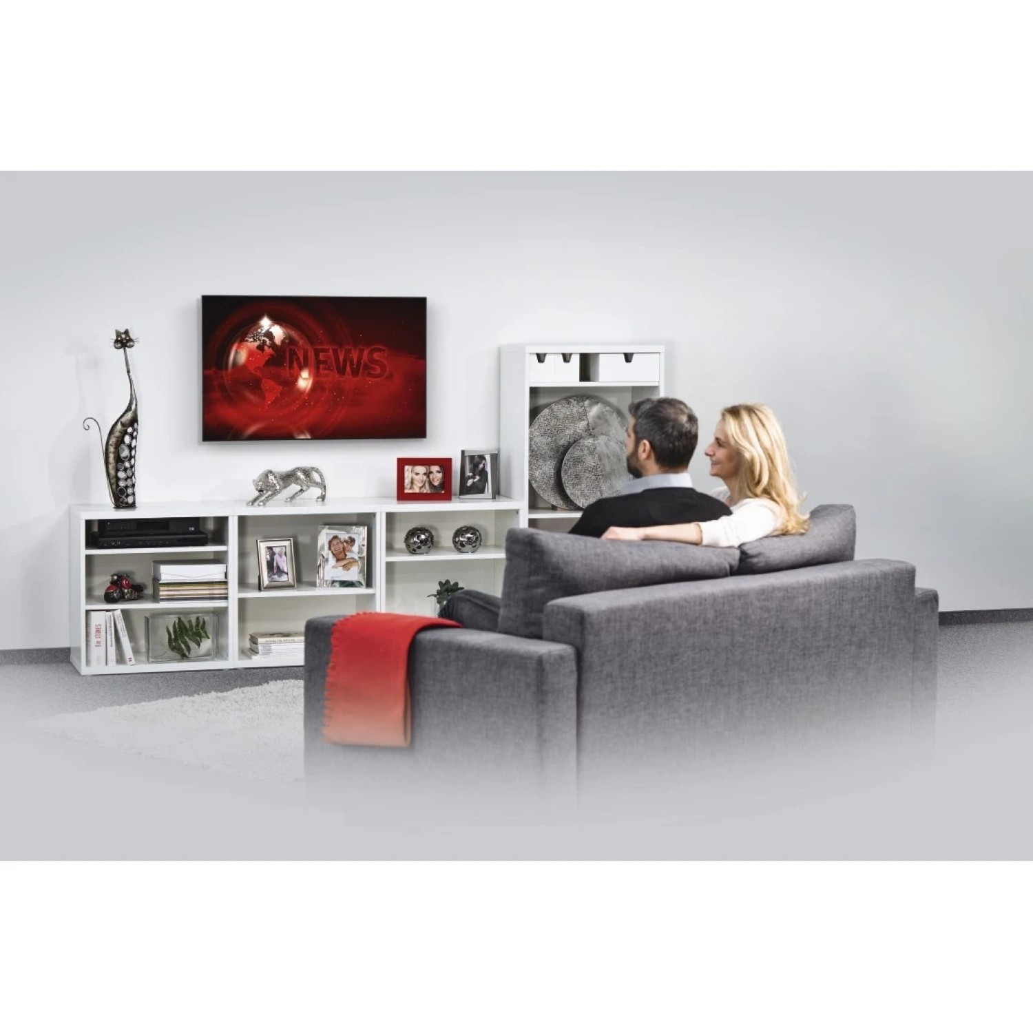 Ein Wohnzimmer mit einem Fernseher über einem weißen Schrank, mit einem Paar auf dem Sofa.