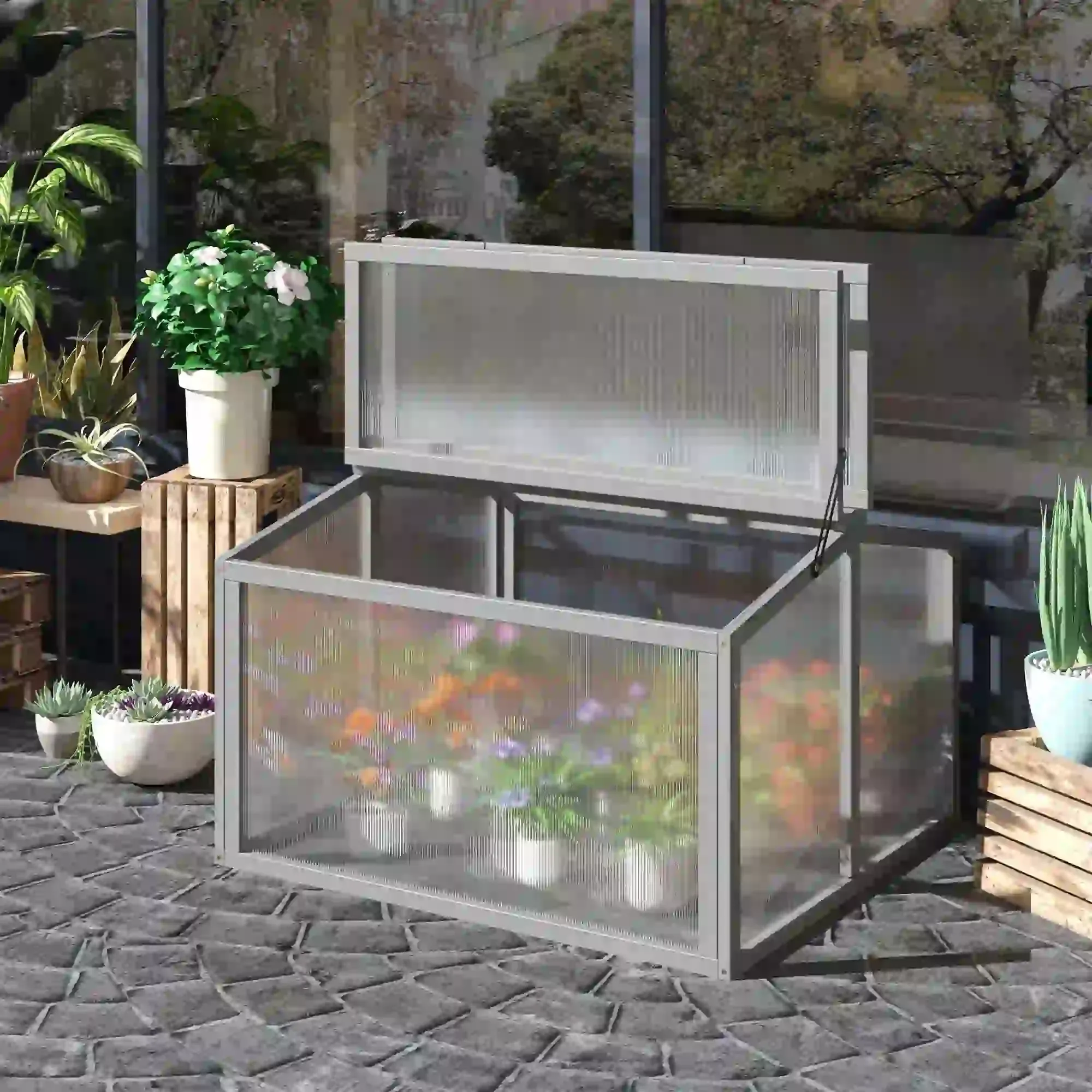 Ein Frühbeetkasten mit klarem Deckel ist geöffnet und gibt den Blick auf Blumen im Inneren frei. Weitere Pflanzen befinden sich in der Umgebung.