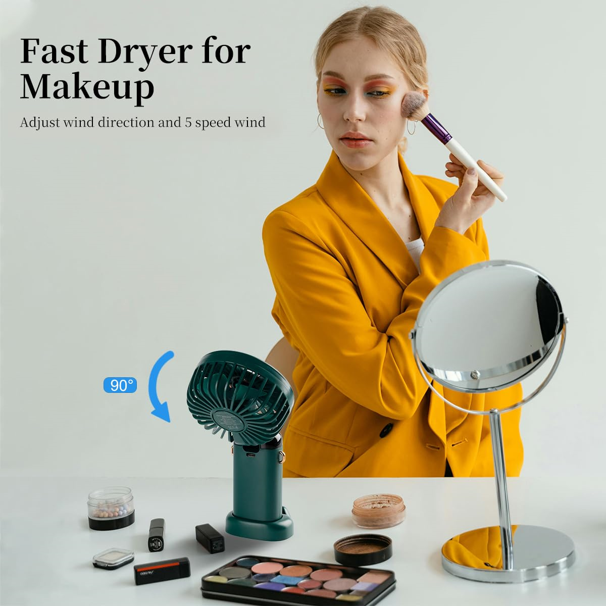 Eine Frau in einer gelben Jacke trägt mit einem Pinsel Make-up auf, mit einem Ventilator, Spiegel und Make-up auf dem Tisch.