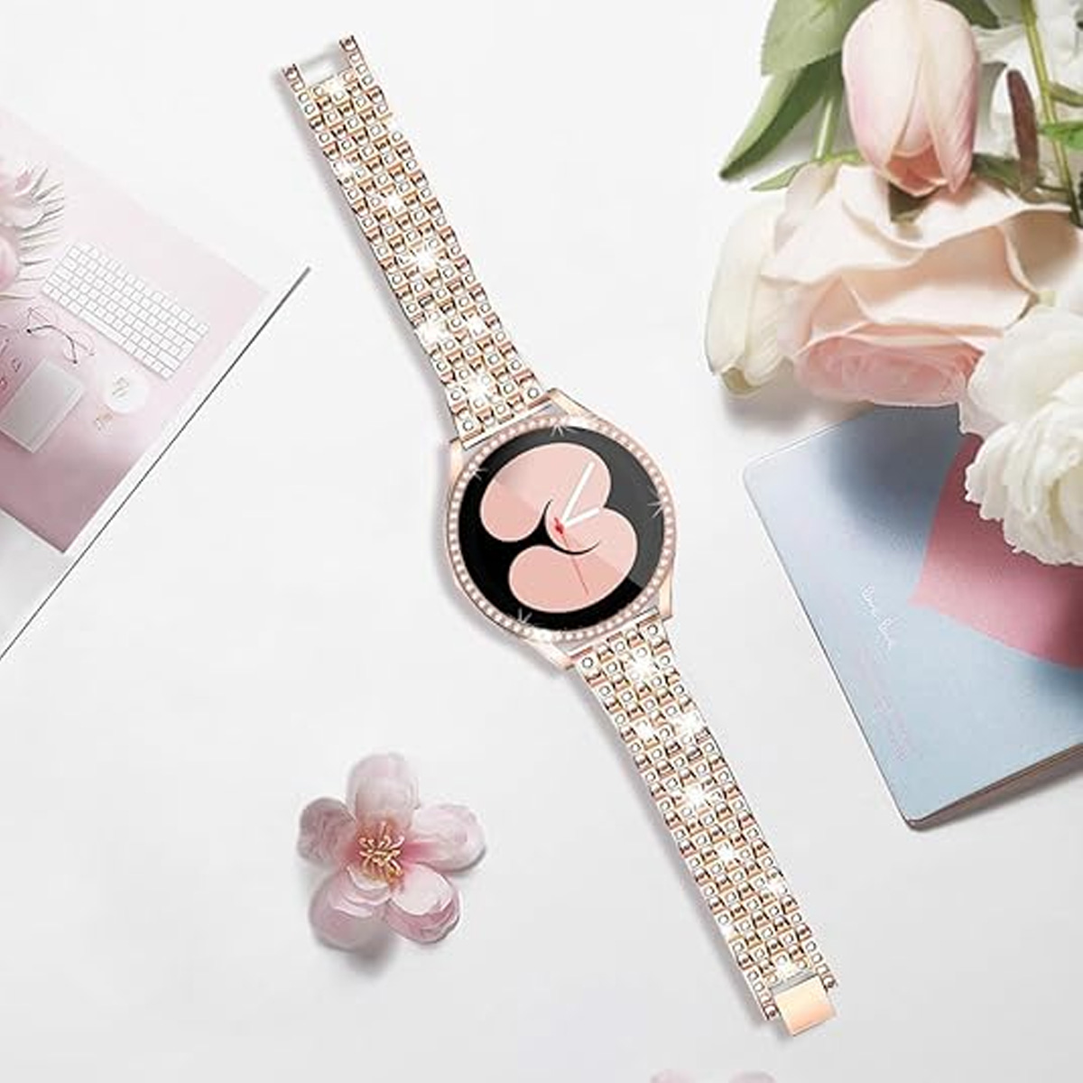 Eine roségoldene Uhr mit einem juwelenbesetzten Armband wird zusammen mit Blumen und einem Notizbuch auf einem weißen Tisch ausgestellt.