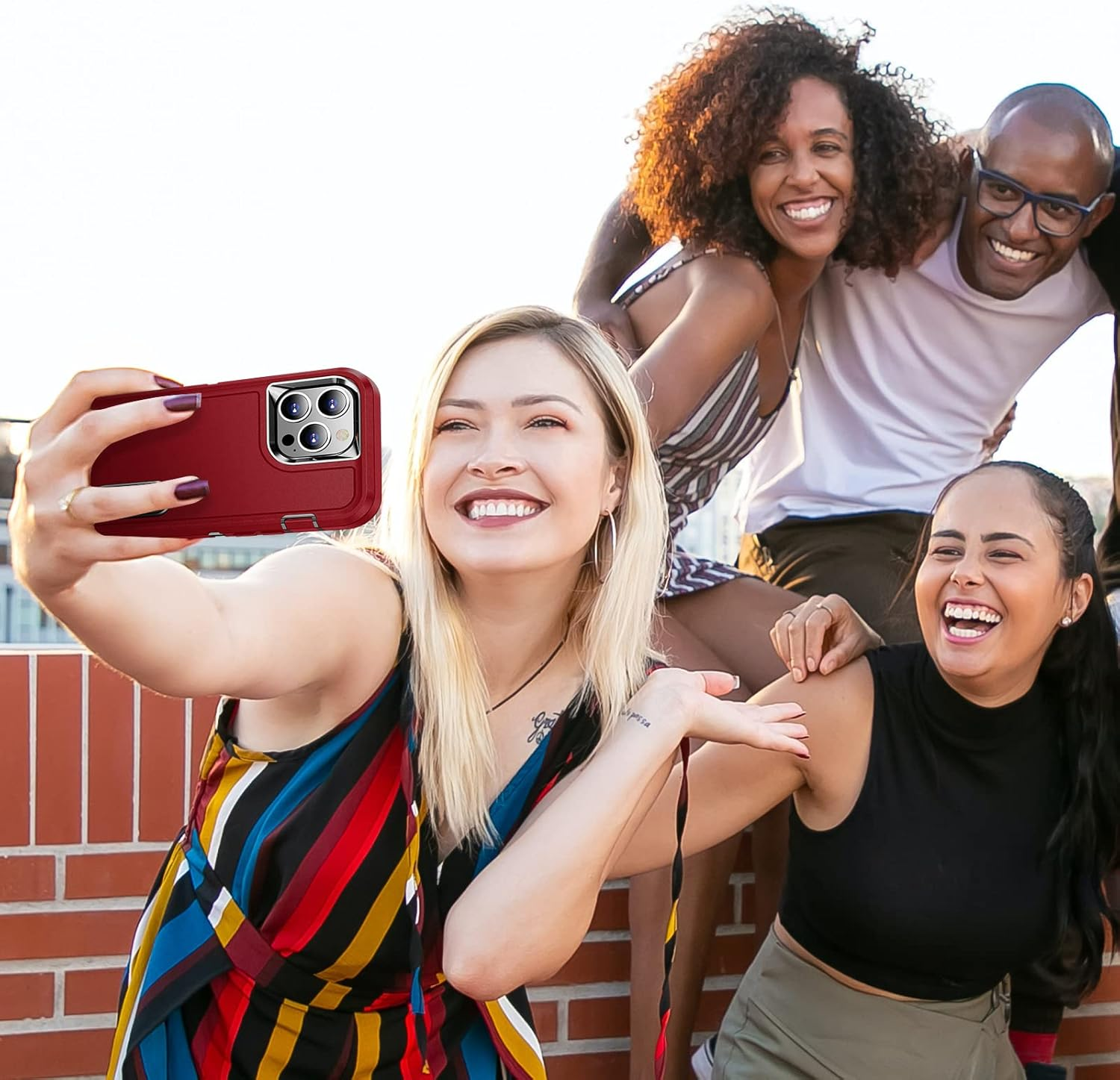 Vier lächelnde Personen posieren für ein Selfie mit einer roten Handyhülle. Sie stehen auf einer Backsteinoberfläche vor einem hellen Himmel.