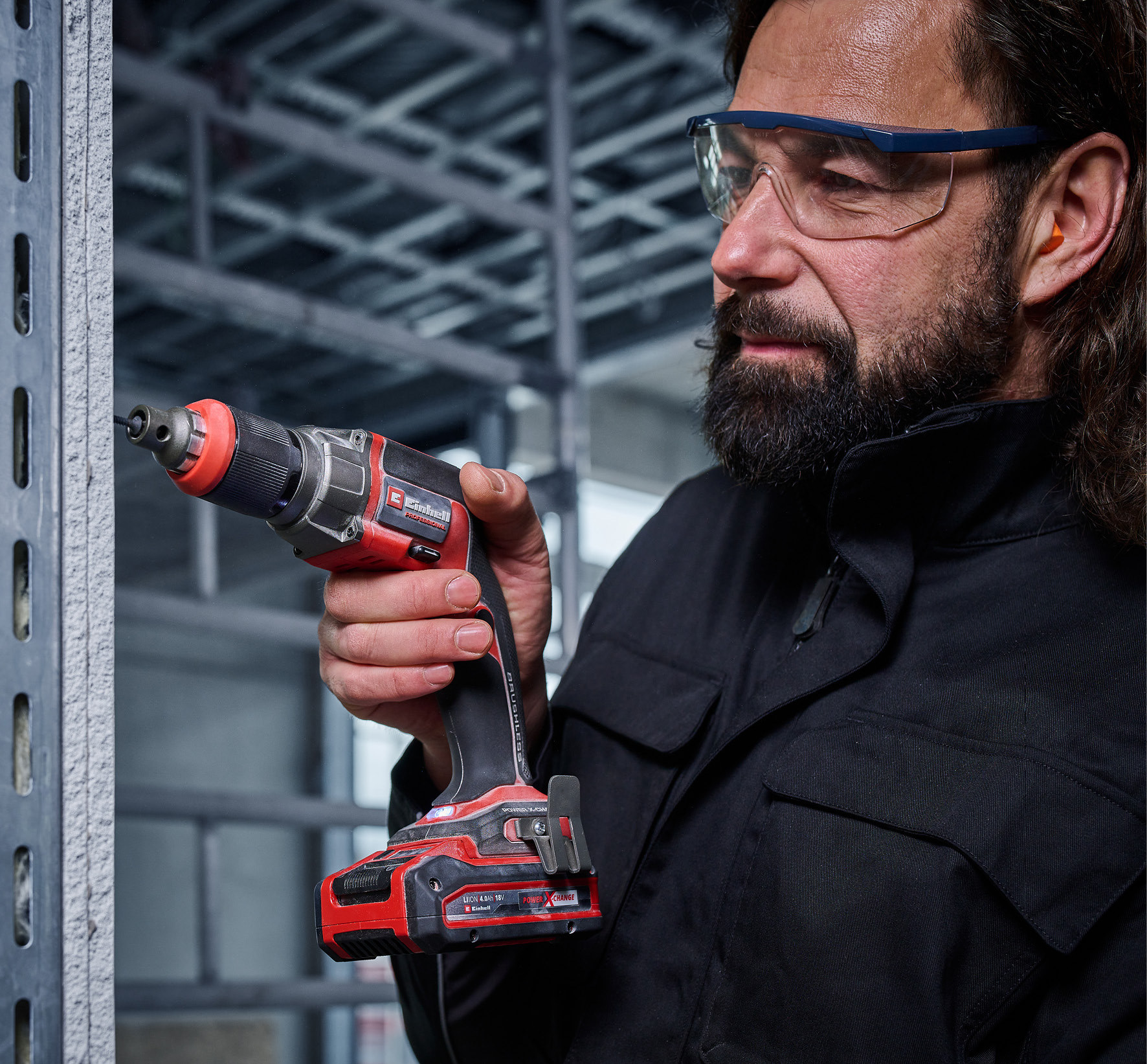 Ein Mann mit Schutzbrille benutzt in einer Baustelle einen rot-schwarzen Einhell Trockenbauschrauber an einer grauen Wand.