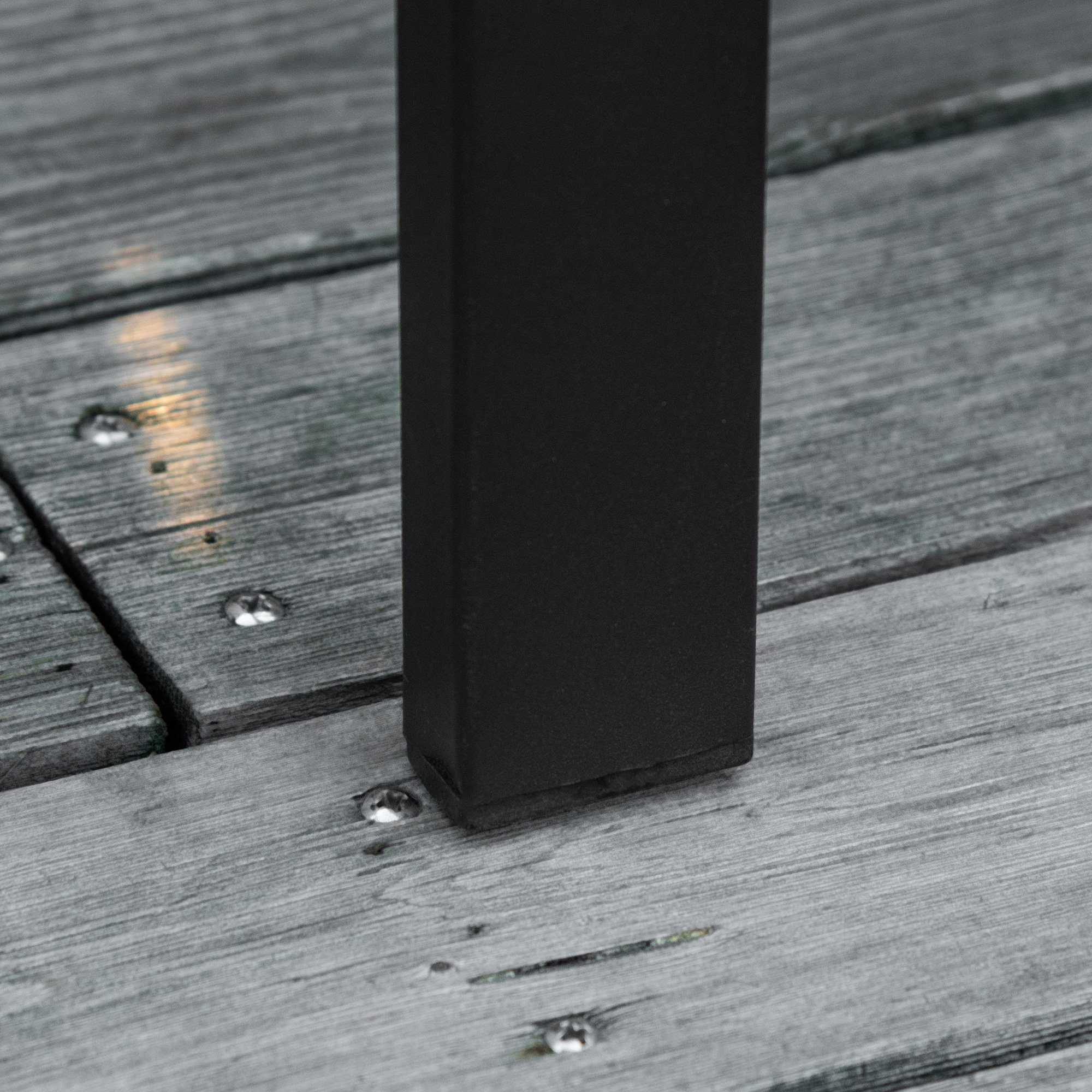 Una pata de silla de metal negro se apoya sobre una plataforma de madera gris, mostrando textura y detalle.