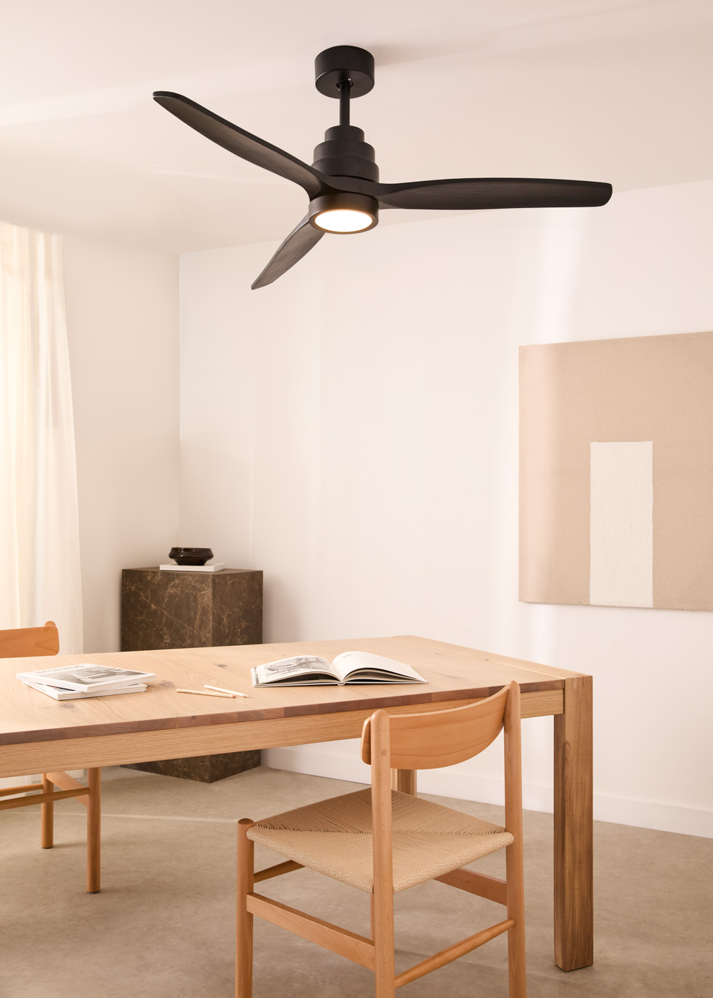 Un ventilador de techo negro con tres aspas y una lámpara en una habitación blanca sobre una mesa y sillas de madera.