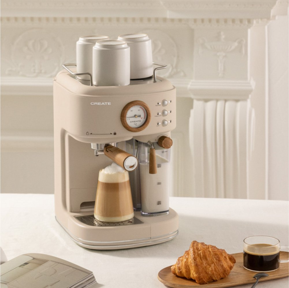 Una máquina de café espresso CREATE beige dispensa café en un vaso sobre una mesa blanca, con croissants y tazas cerca.