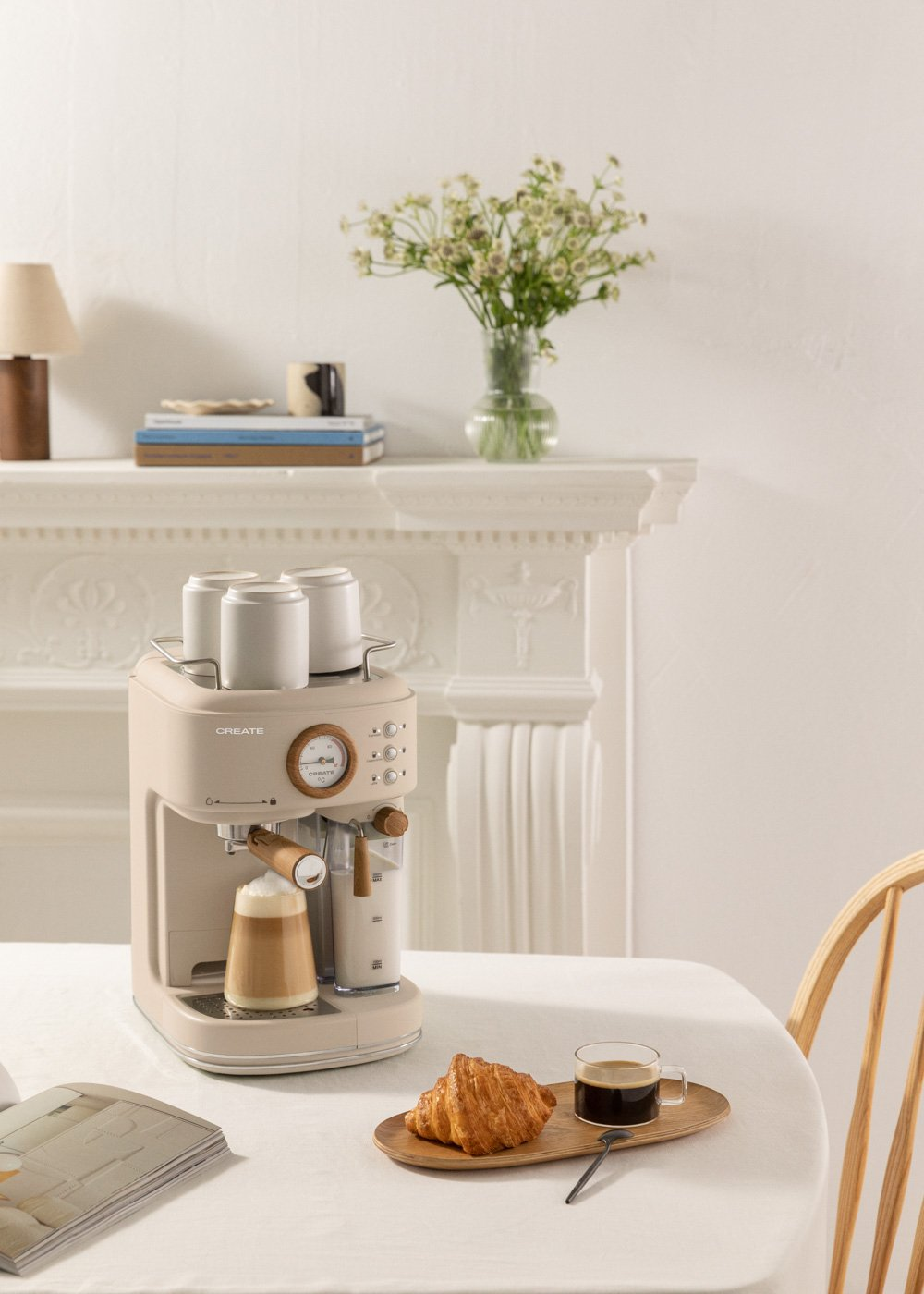 Una cafetera beige con un vaso de café está sobre una mesa. Un cruasán y una taza de café también están sobre la mesa.