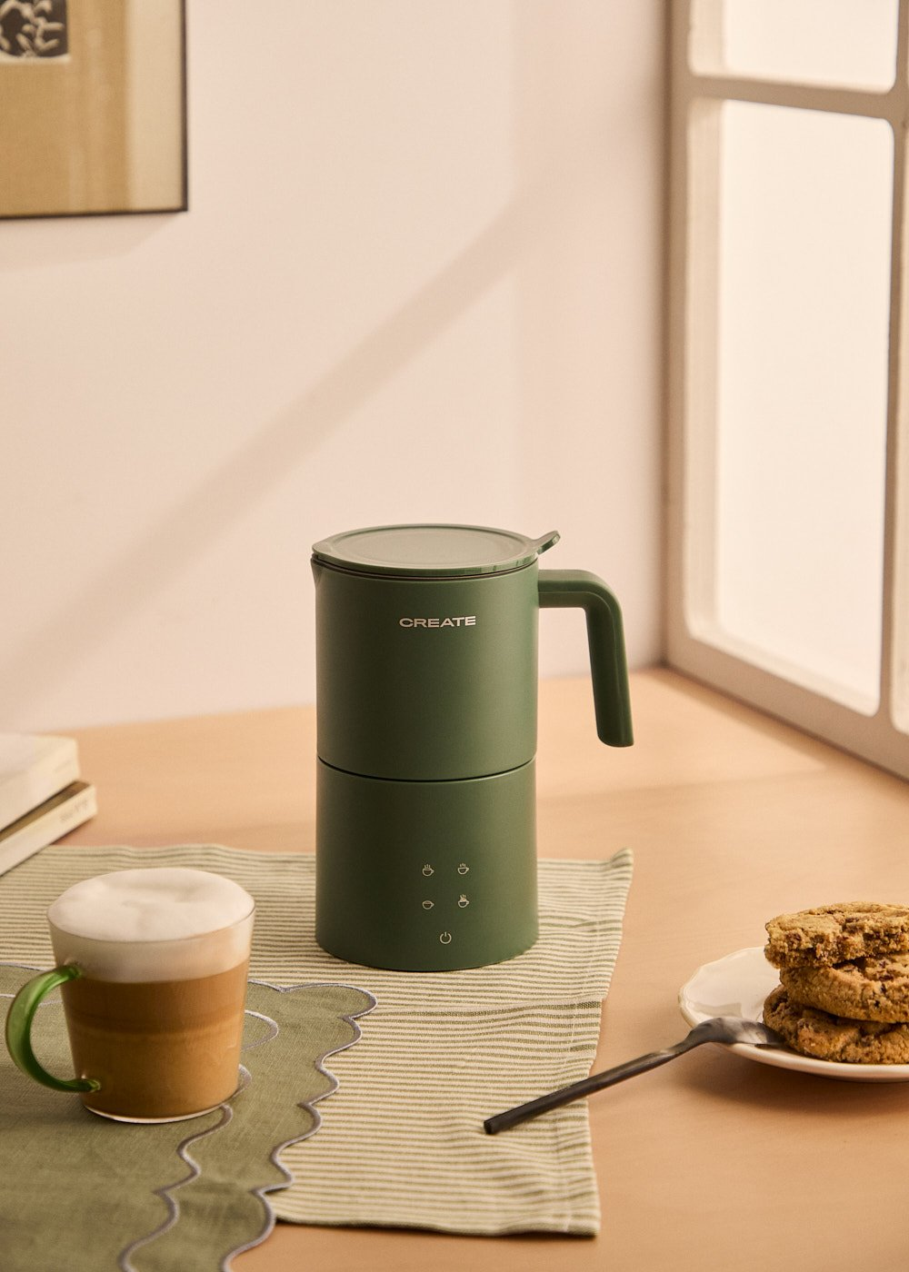 Un espumador de leche verde con una taza de café y galletas están en la mesa. Una ventana está en el fondo.
