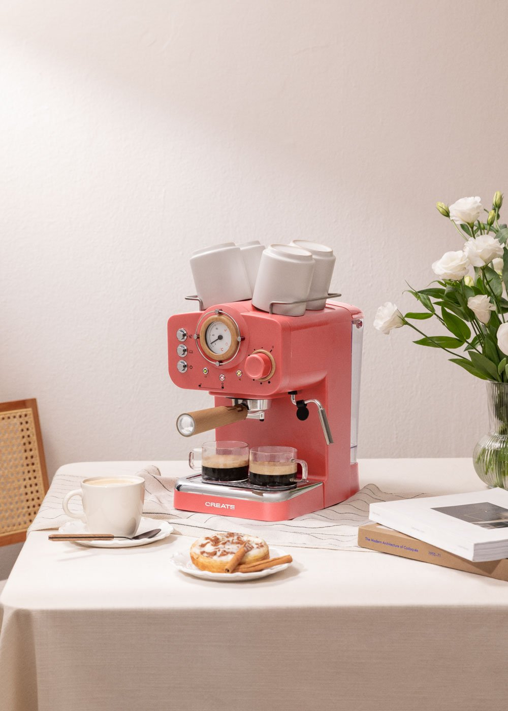 Una máquina de café espresso rosa se encuentra sobre una mesa con tazas, café y un pastel.