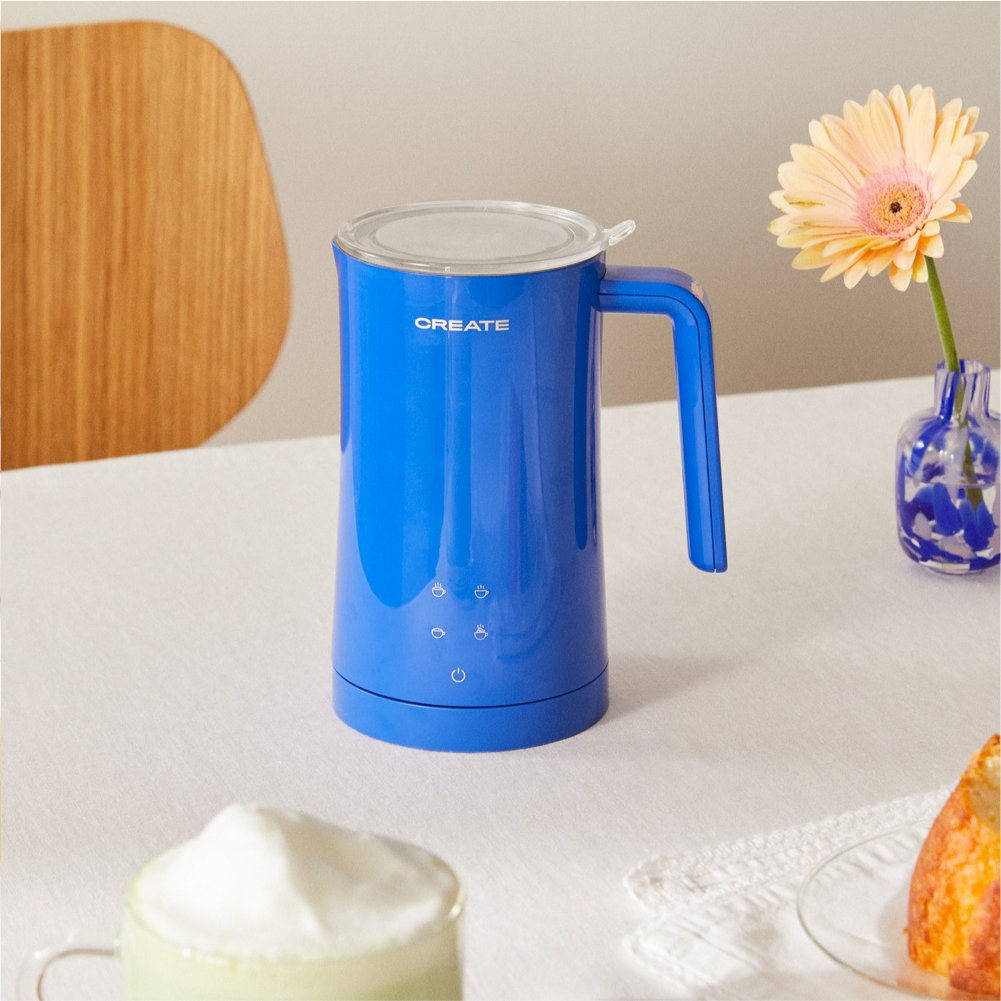 Un espumador de leche azul Create se sienta en una mesa con una flor, una bebida y un pastel.