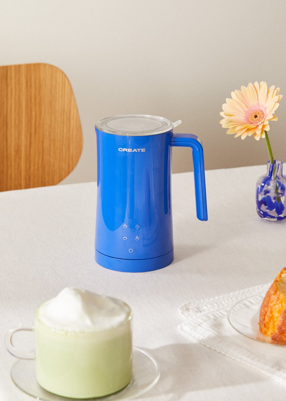 Un espumador de leche azul Create está sobre una mesa con una bebida, un pastel y una flor.