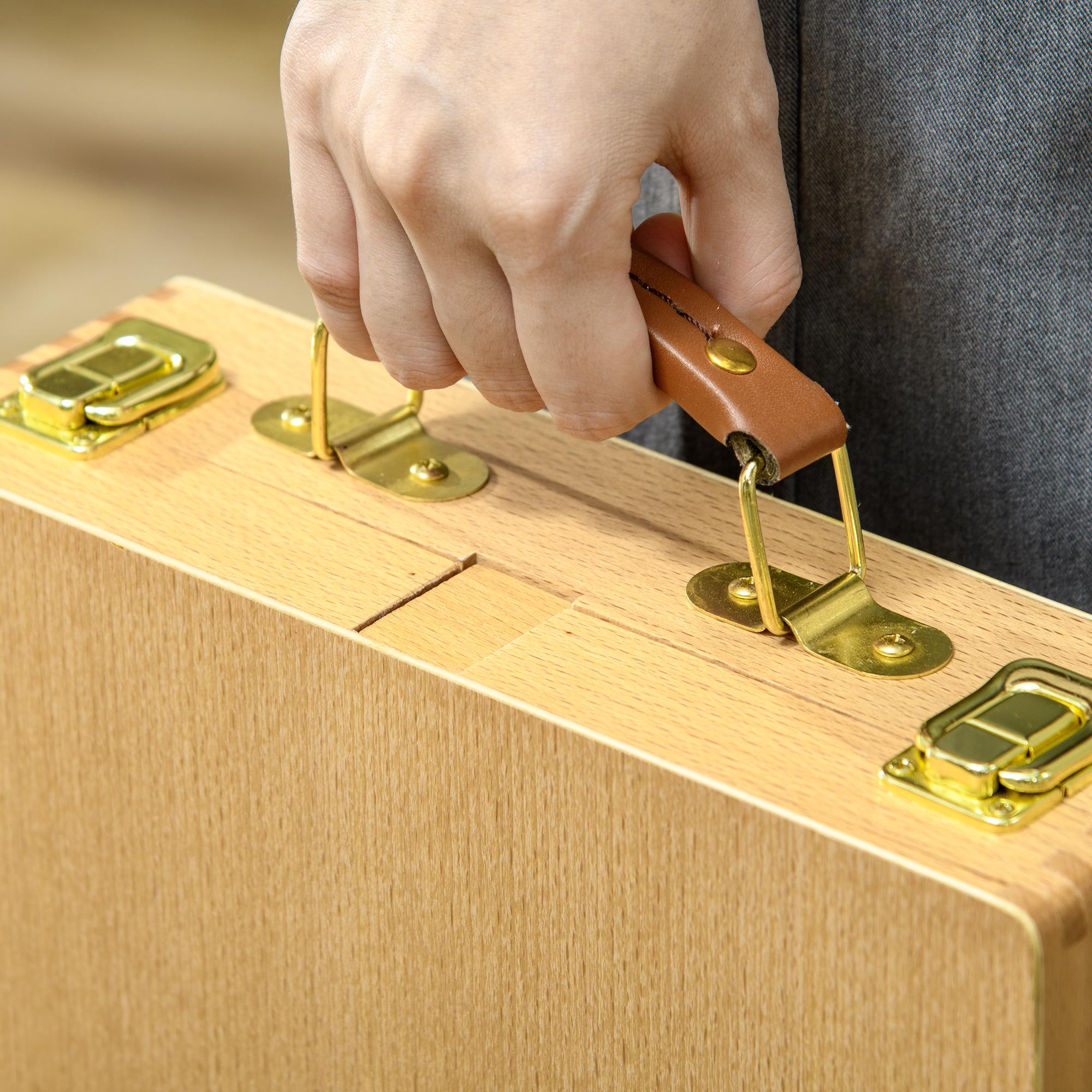 Eine Hand greift den braunen Ledergriff einer hellen Holzkiste mit goldenen Verschlüssen und Beschlägen.
