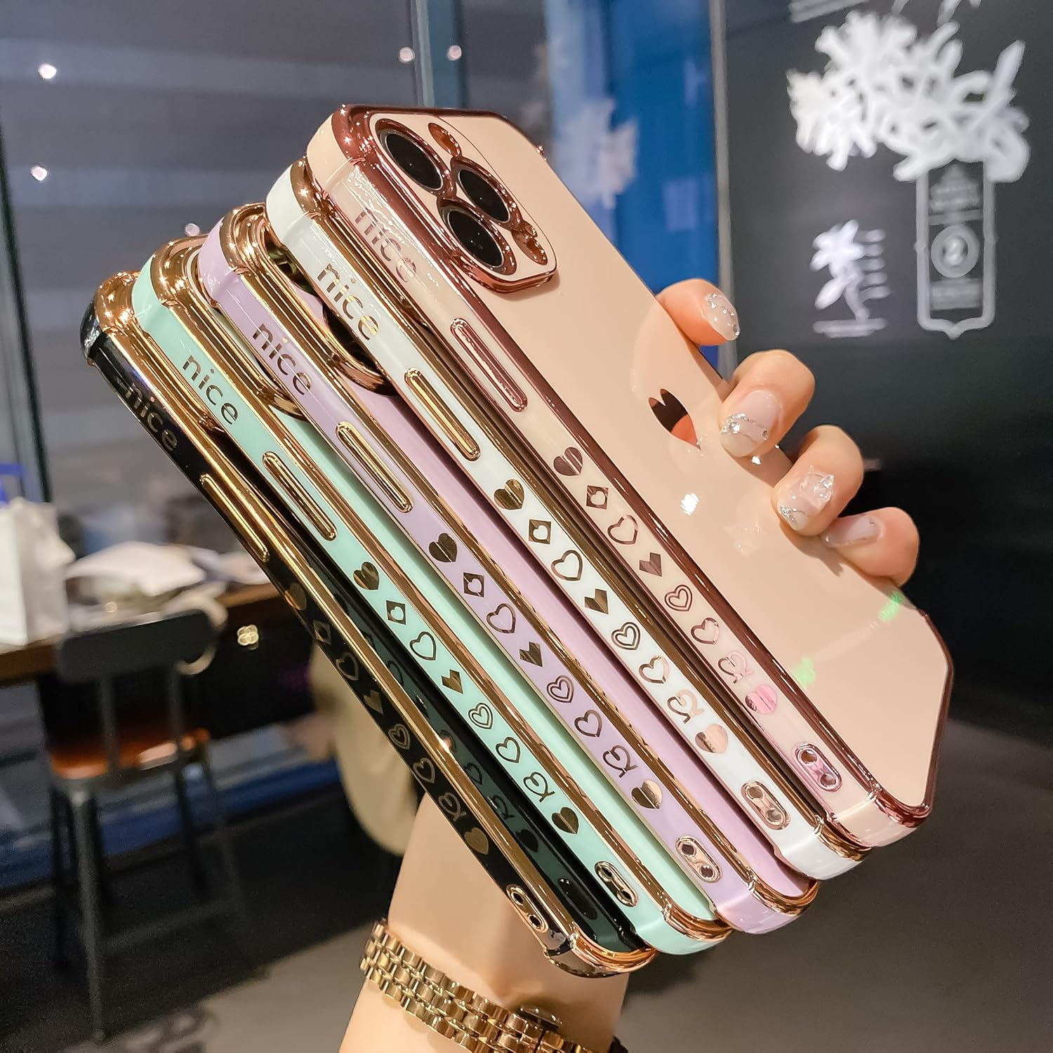 Hand hält einen Stapel farbenfroher iPhone-Hüllen mit goldenen Akzenten und Herzmotiven. Die Hüllen sind schwarz, grün, rosa und weiß.