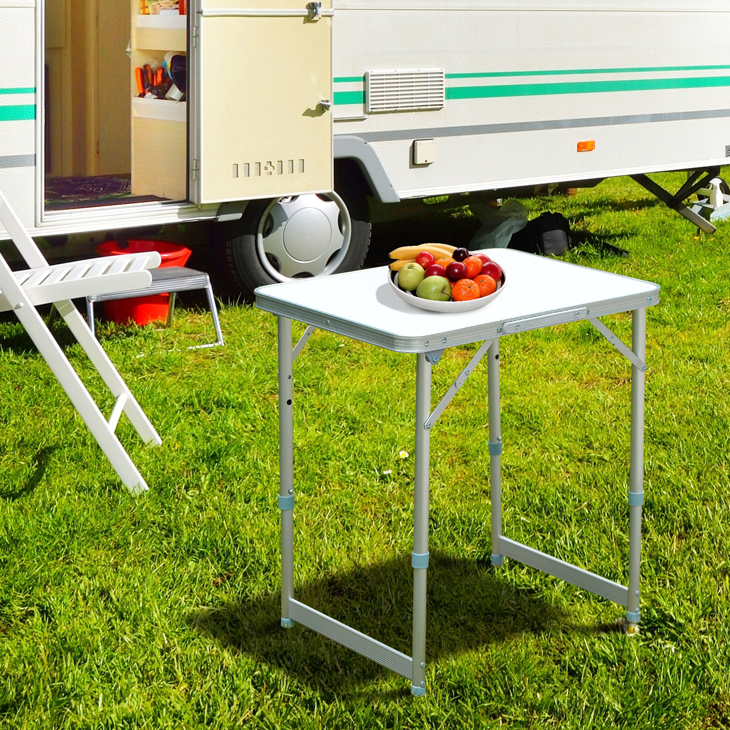 Una mesa plegable blanca con fruta encima está colocada cerca de una caravana en un campo de hierba.