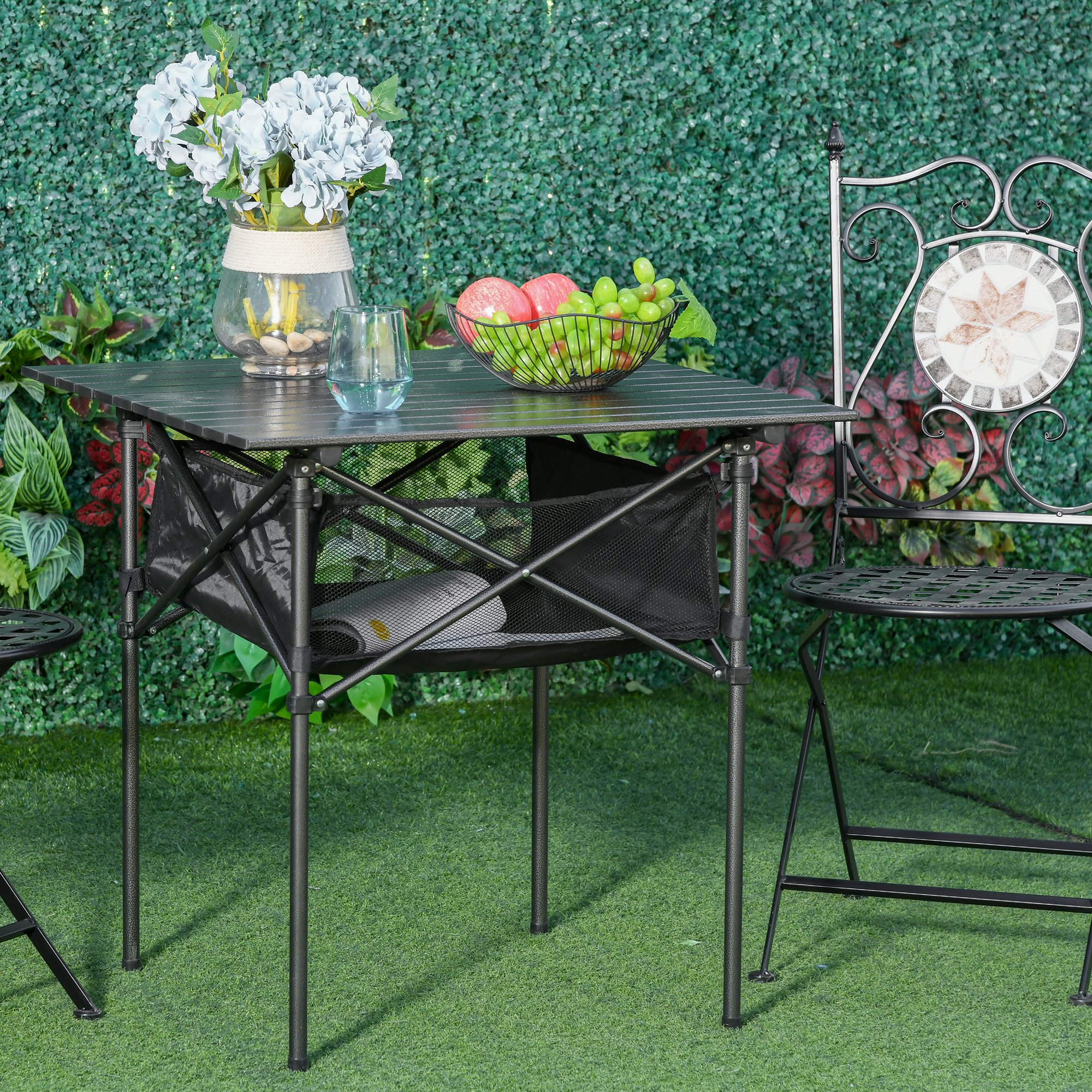 Una mesa plegable negra con un área de almacenamiento de malla, un jarrón, un frutero y una silla, sobre hierba verde contra una pared verde.