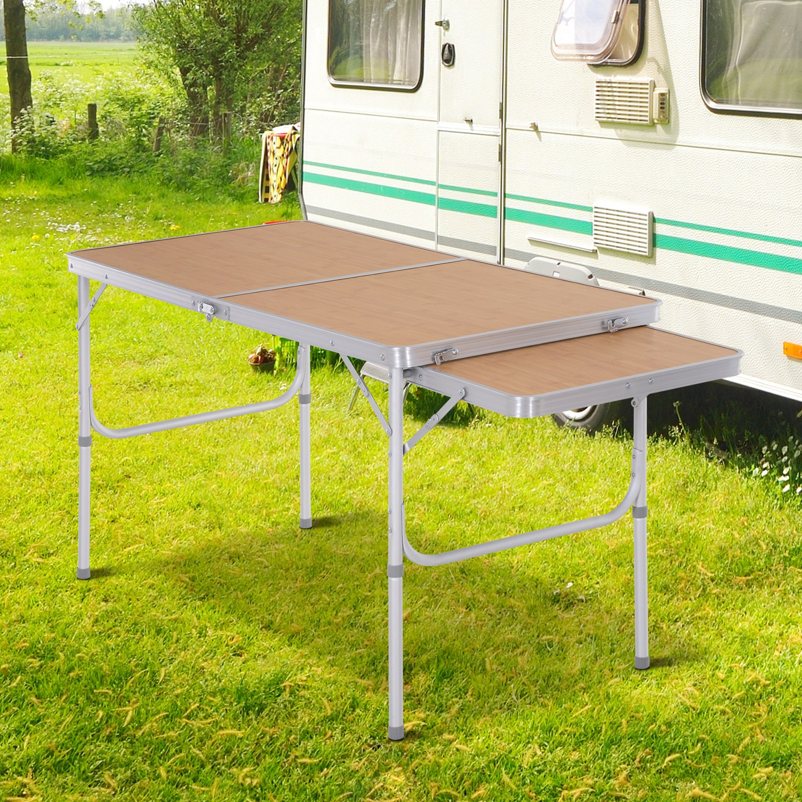 Una mesa plegable con una tapa de madera clara y un marco plateado está instalada afuera cerca de una caravana.