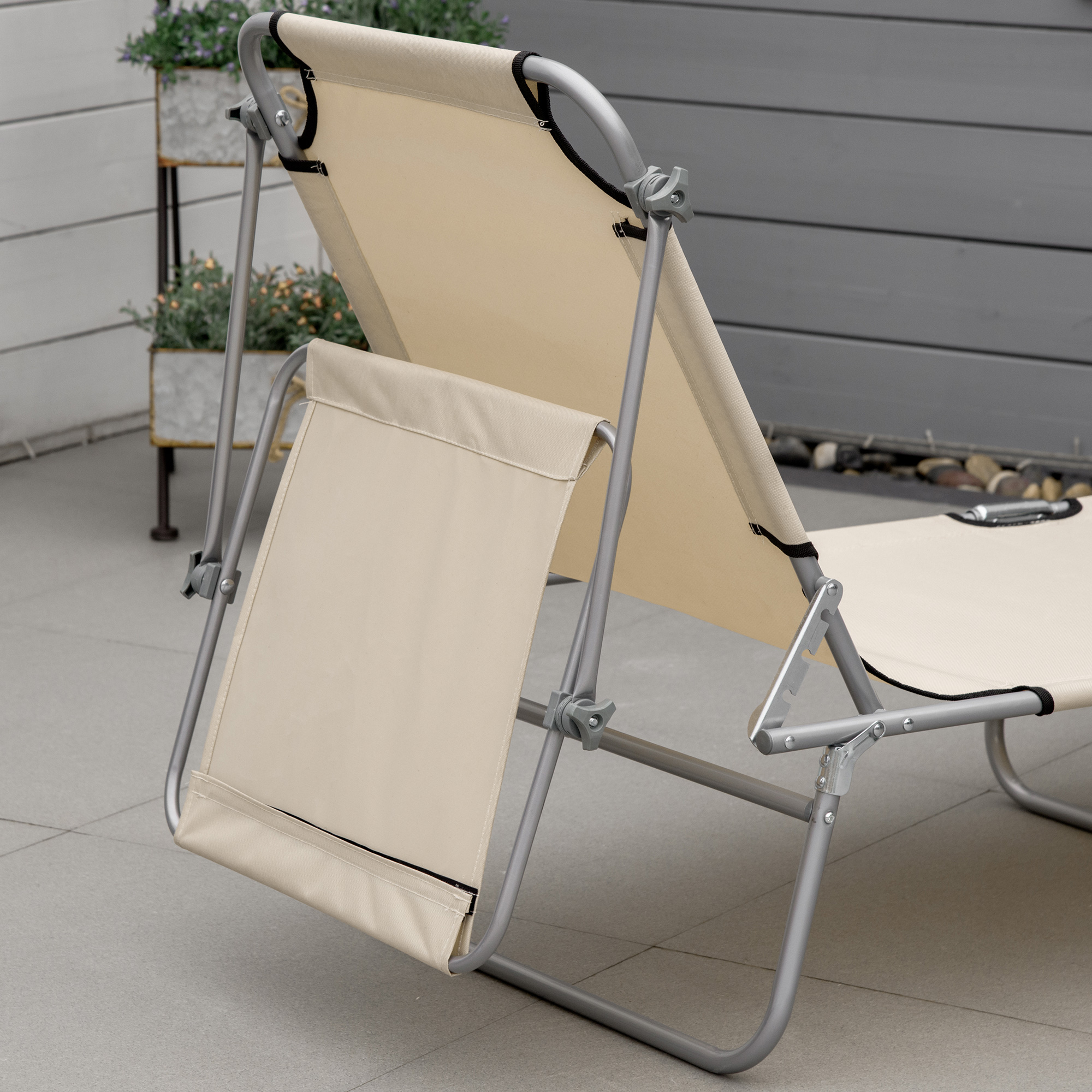 Una sedia a sdraio beige con struttura in metallo è posizionata su una superficie piastrellata, con piante sullo sfondo.