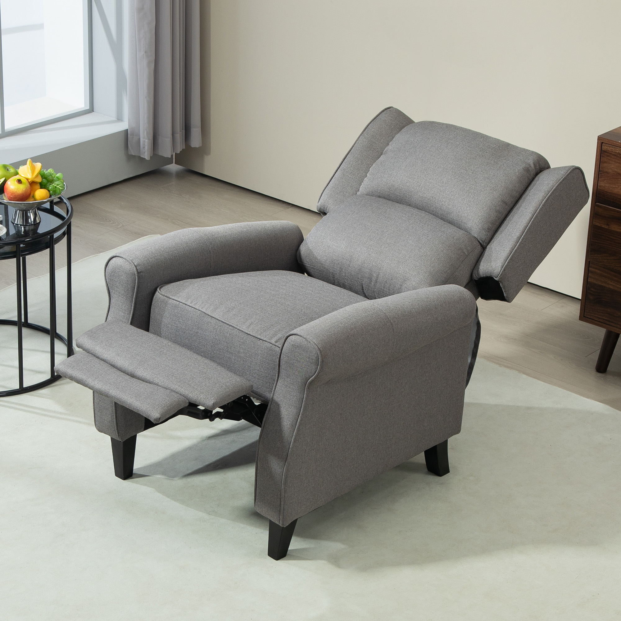 Sillón gris en la habitación. Posición reclinada, reposapiés arriba. Mesa auxiliar con fruta, alfombra blanca.