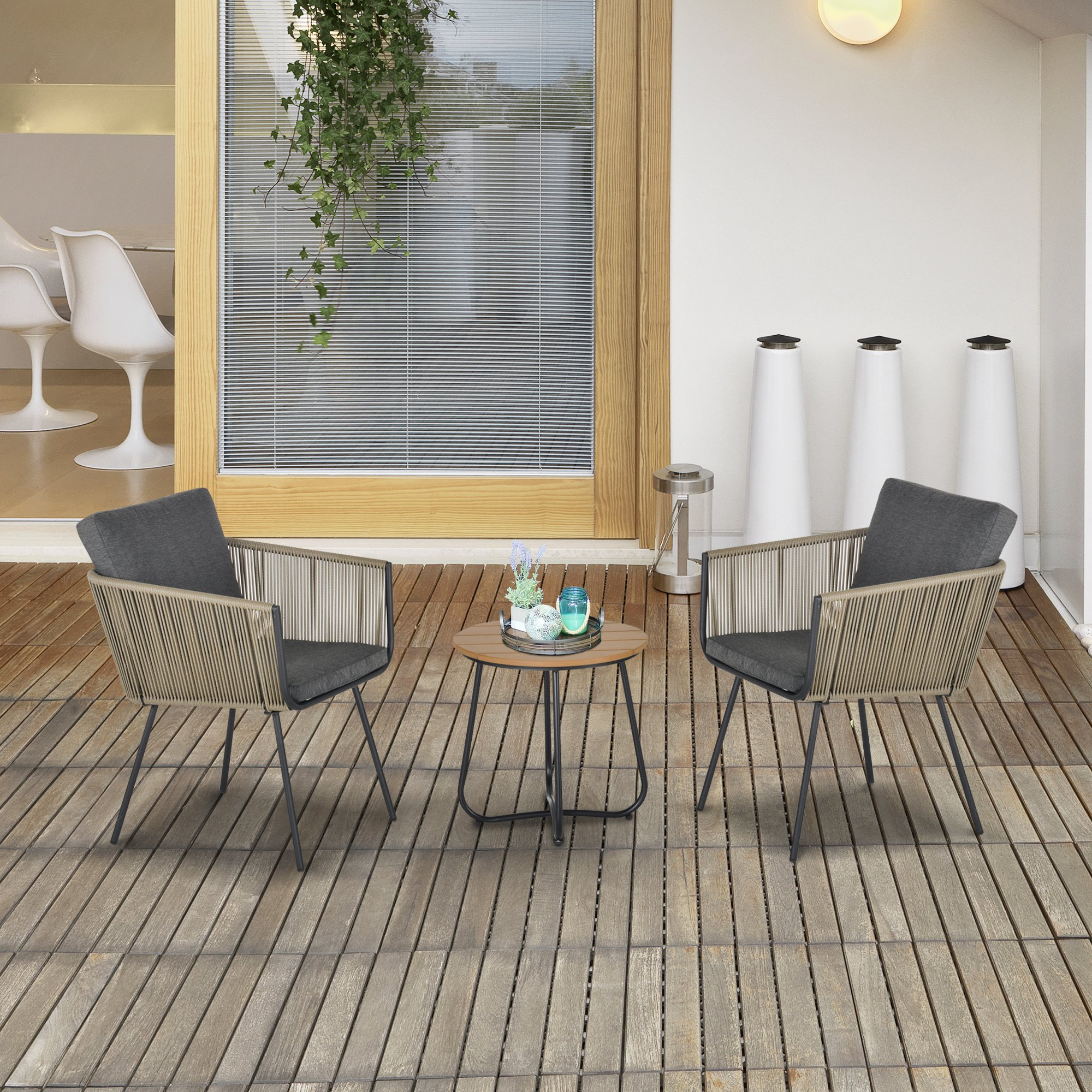Dos sillas de cuerda y una mesa se sientan en una terraza de madera con una puerta y jarrones blancos al fondo.