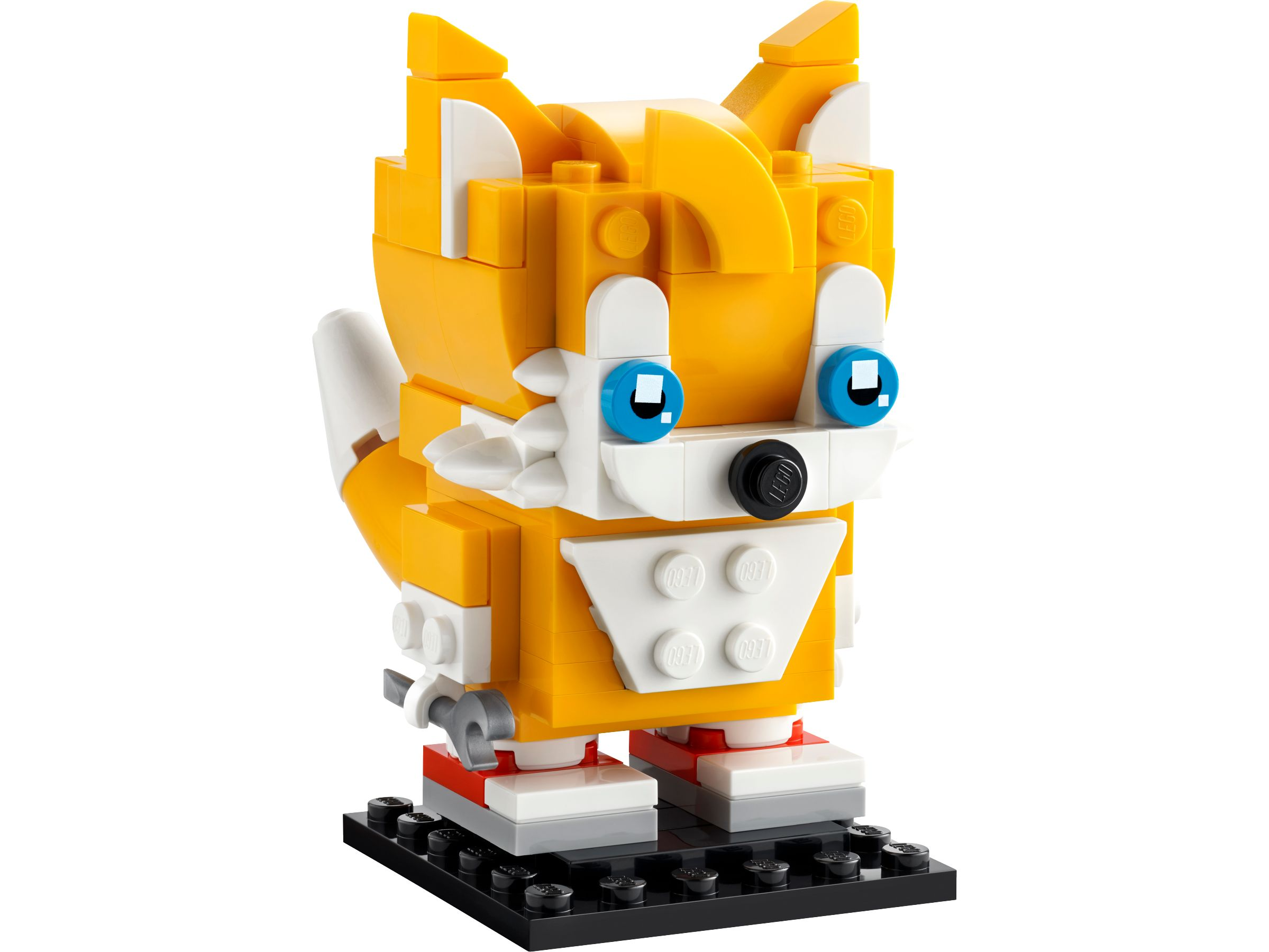 Eine LEGO Tails Figur, orange und weiß, mit blauen Augen, auf schwarzer Basis, weißer Hintergrund.