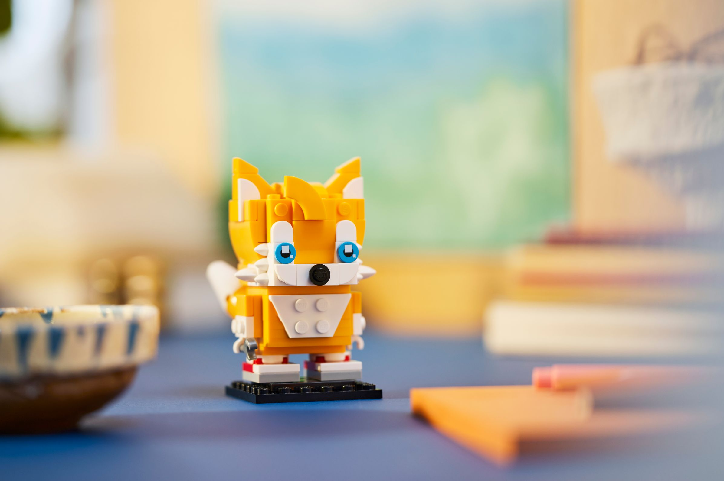 Eine LEGO Tails Figur auf einer schwarzen Basis, mit verschwommenem Hintergrund von Schreibtisch und Objekten.