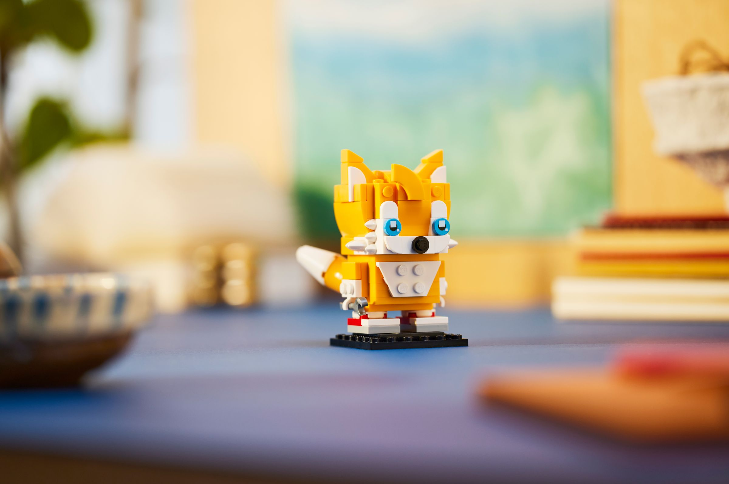 Eine LEGO Tails Figur auf einer schwarzen Basis, mit verschwommenem Hintergrund von Schreibtisch und Objekten.