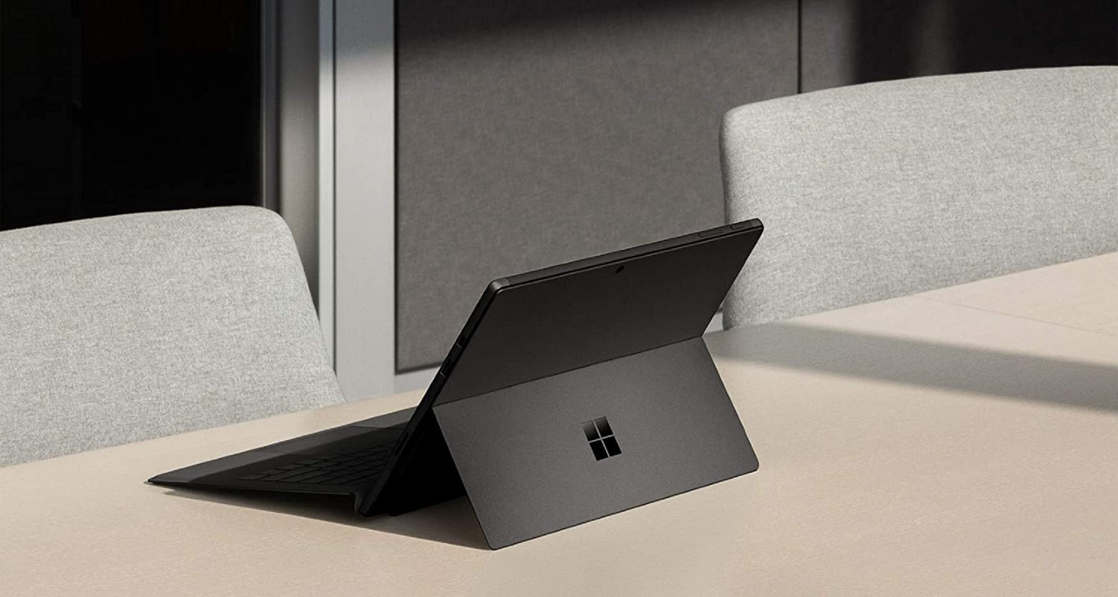 Ein schwarzer Laptop mit Microsoft-Logo, geöffnet auf einer hellen Oberfläche, im Hintergrund zwei Stühle, Büroumgebung.