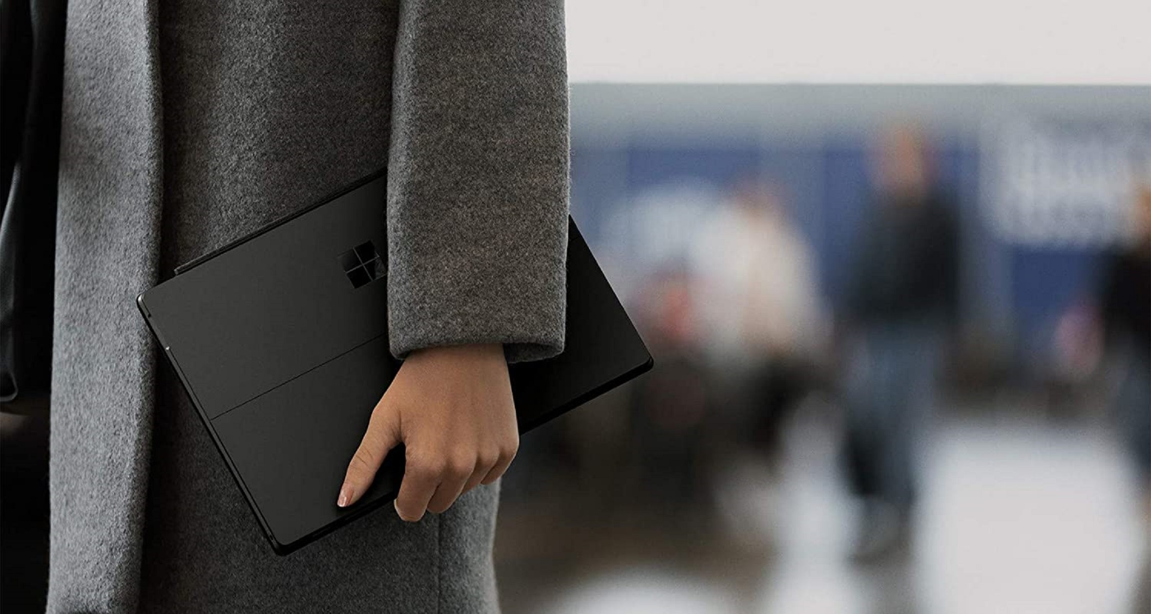 Eine Person hält einen schwarzen Laptop mit Microsoft-Logo, trägt einen grauen Mantel, in einem verschwommenen öffentlichen Raum.