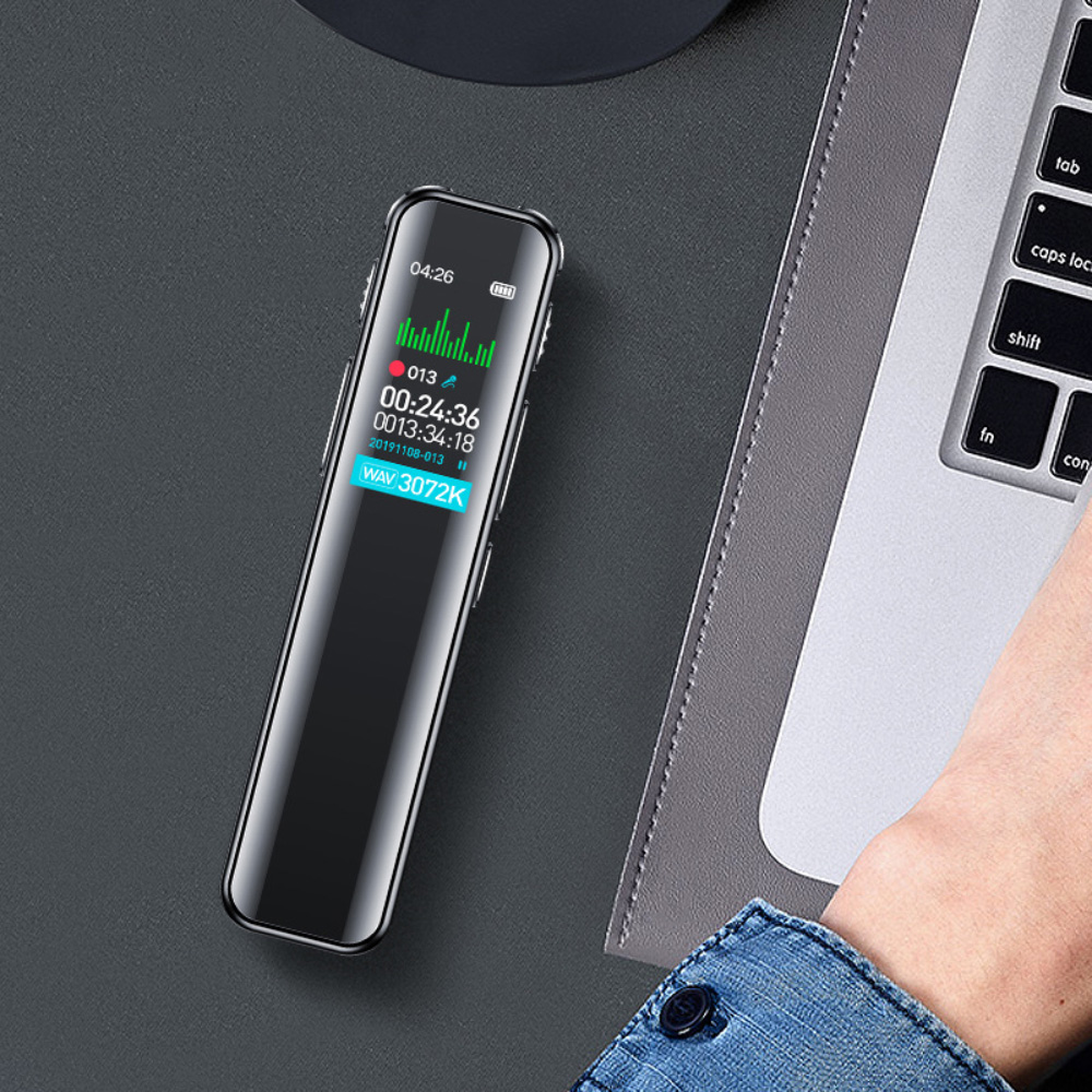 Ein schwarzer Audiorecorder wird auf einer grauen Oberfläche neben einem Laptop platziert, wobei eine Hand und ein Jeansärmel sichtbar sind.