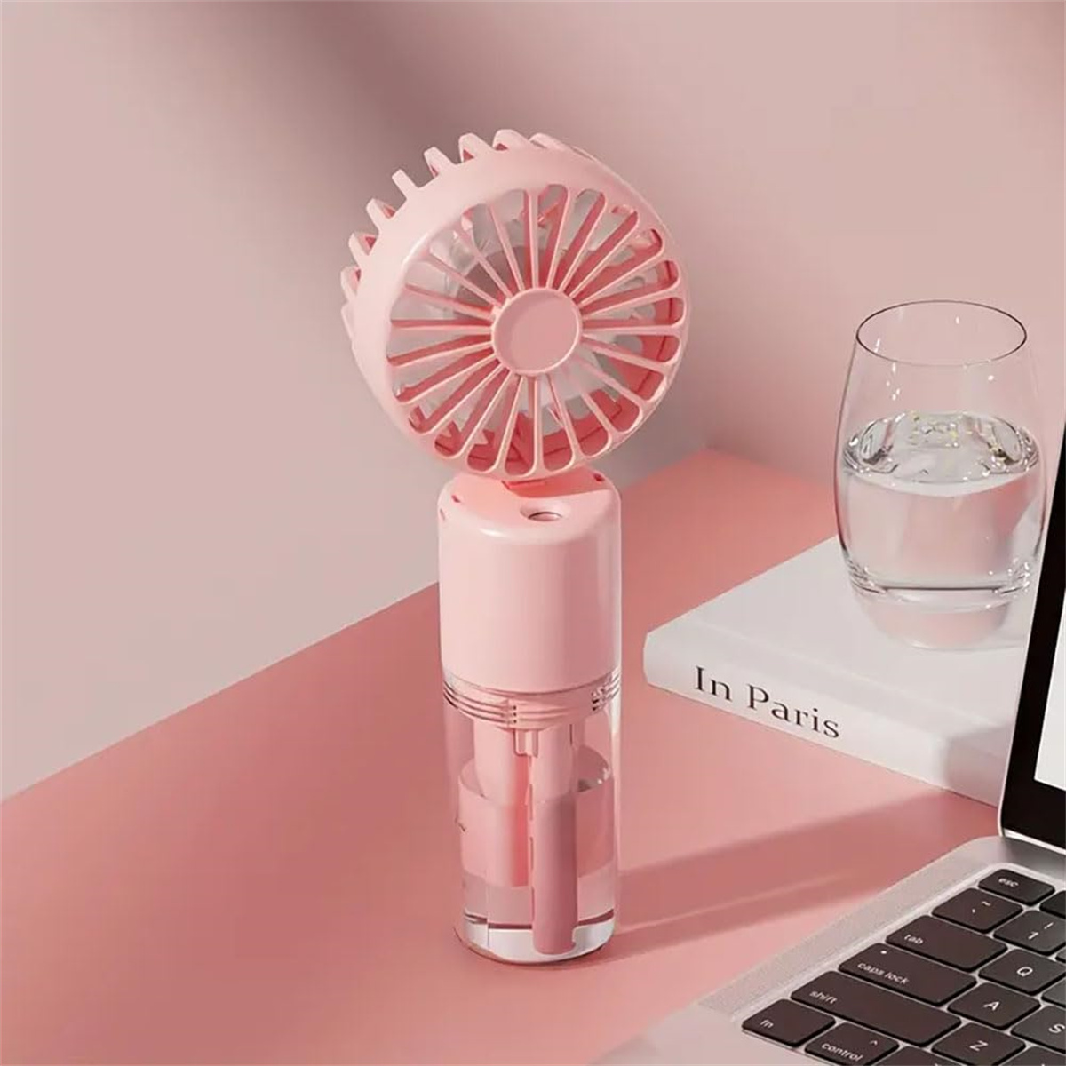 Ein rosa Handventilator mit Wasserbehälter steht auf rosa Oberfläche neben einem Wasserglas.