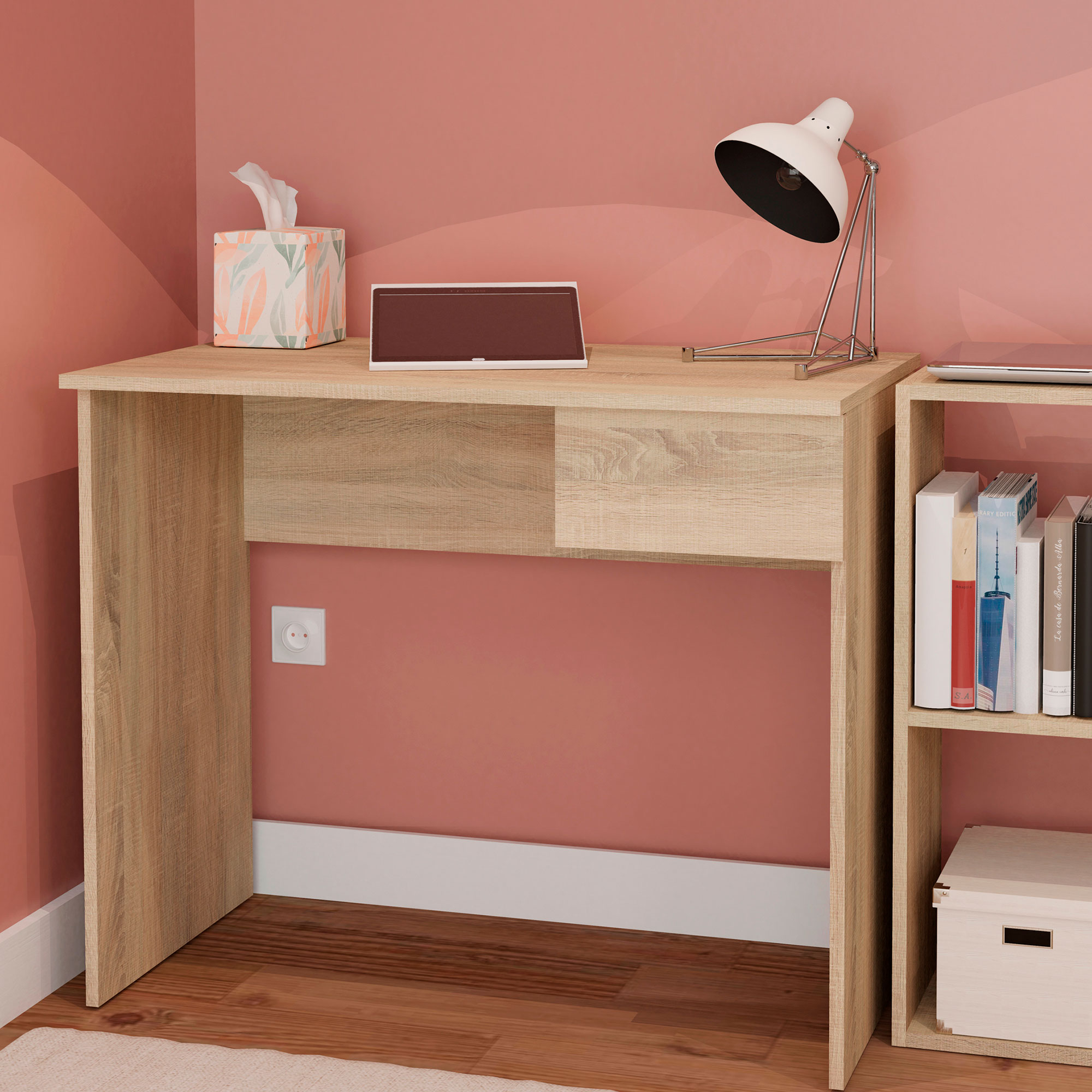Escritorio de madera clara con lámpara, caja de pañuelos y libros. Fondo de pared rosa con enchufe.