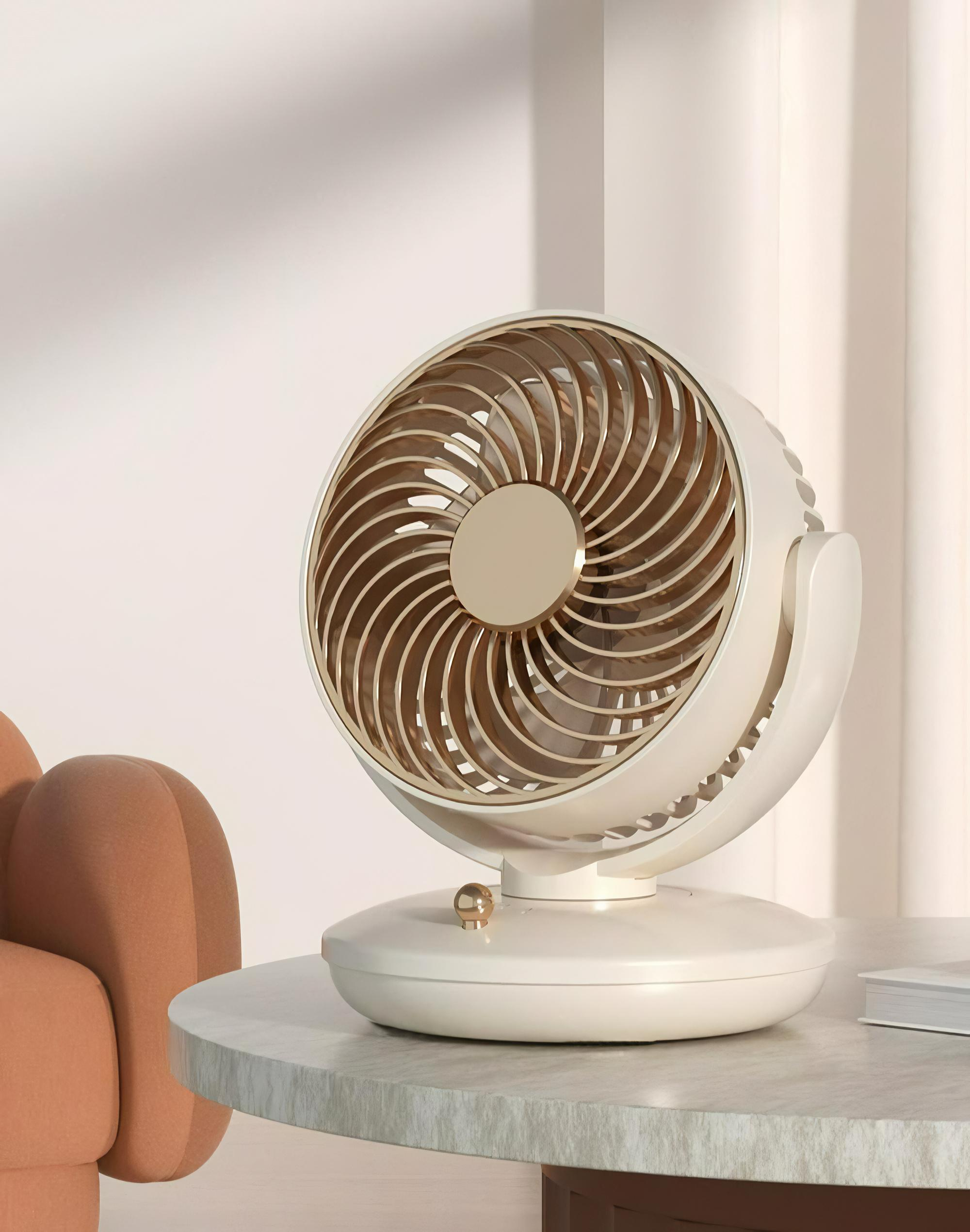 Ein weiß-goldener Ventilator steht auf einem Marmortisch. Im Hintergrund ist ein pfirsichfarbener Stuhl zu sehen.