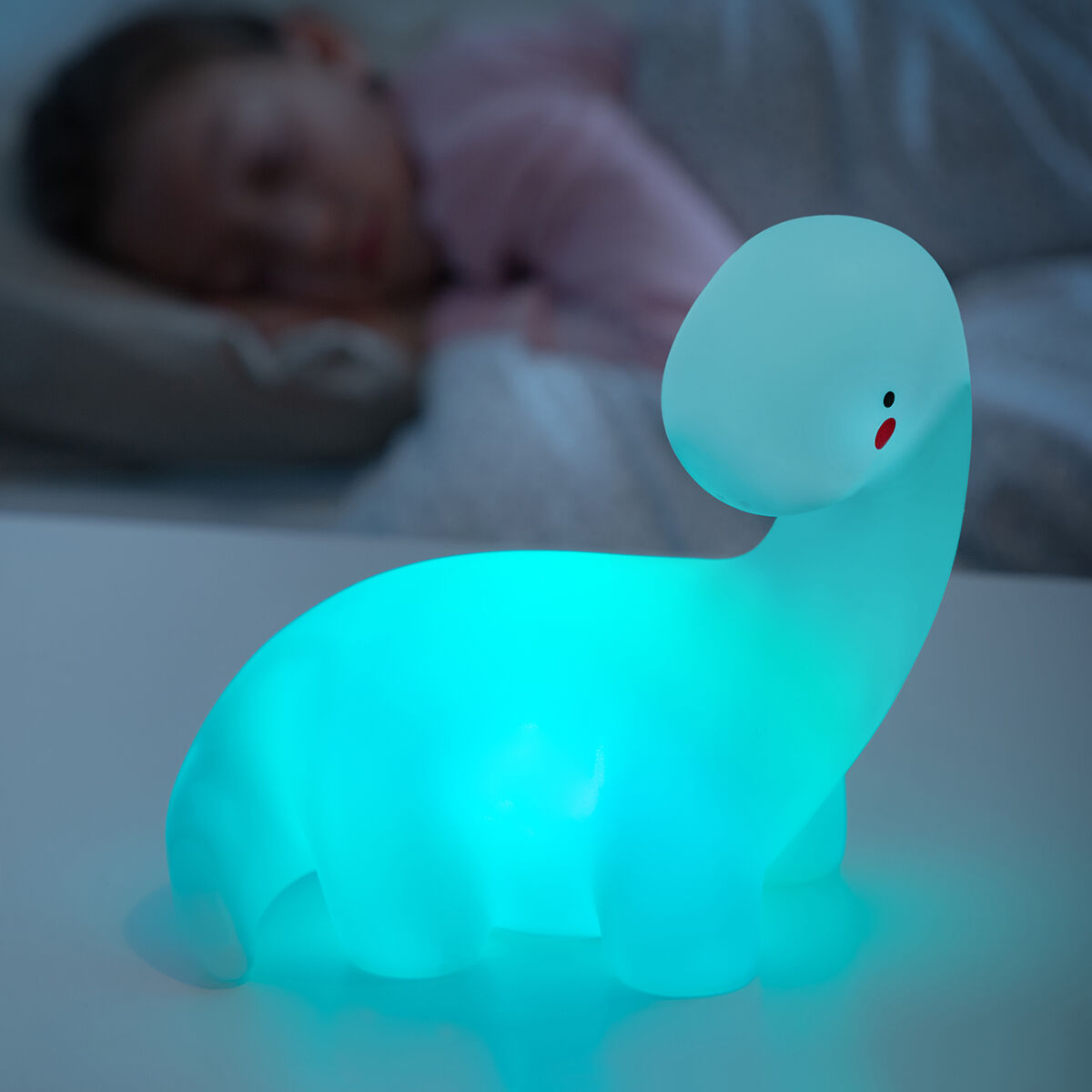 Un bambino che dorme vicino a una luce notturna a dinosauro blu brillante, su una superficie bianca.