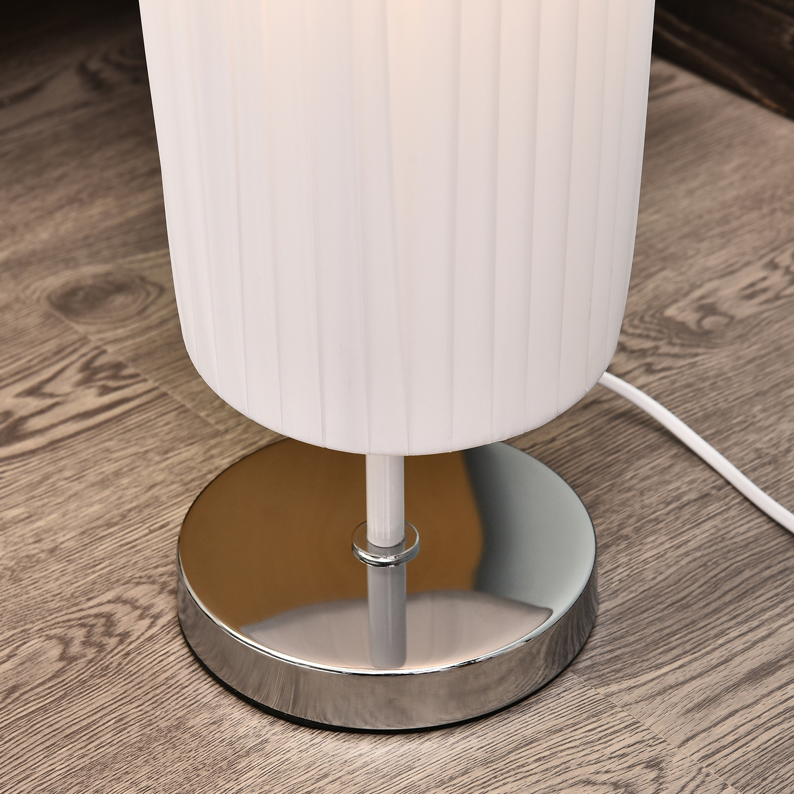 Una lámpara de mesa blanca con una pantalla plisada y una base cromada. Sobre una superficie de madera.