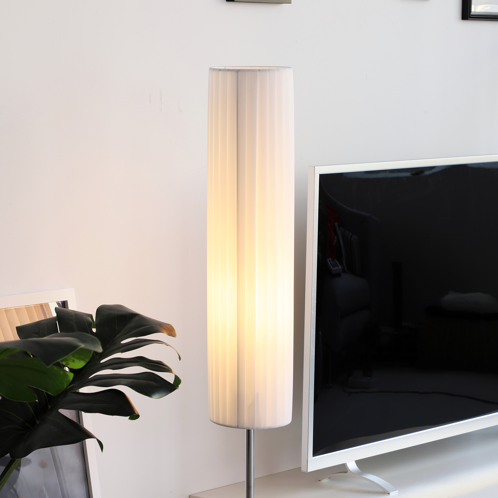 Una lámpara alta blanca con pantalla plisada junto a TV y planta en superficie blanca.