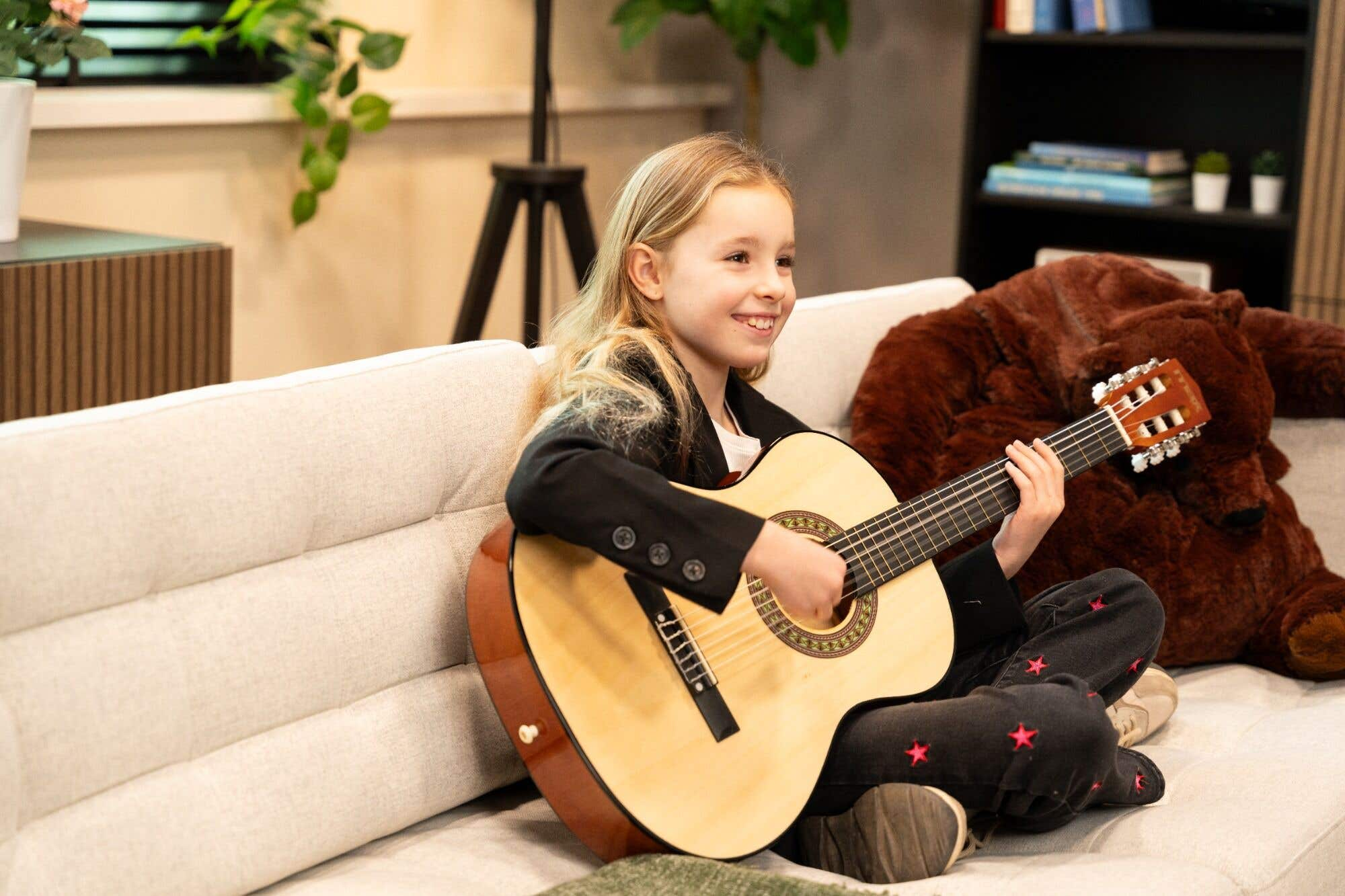 Een jong meisje speelt gitaar terwijl ze op een bank zit, met een teddybeer op de achtergrond.