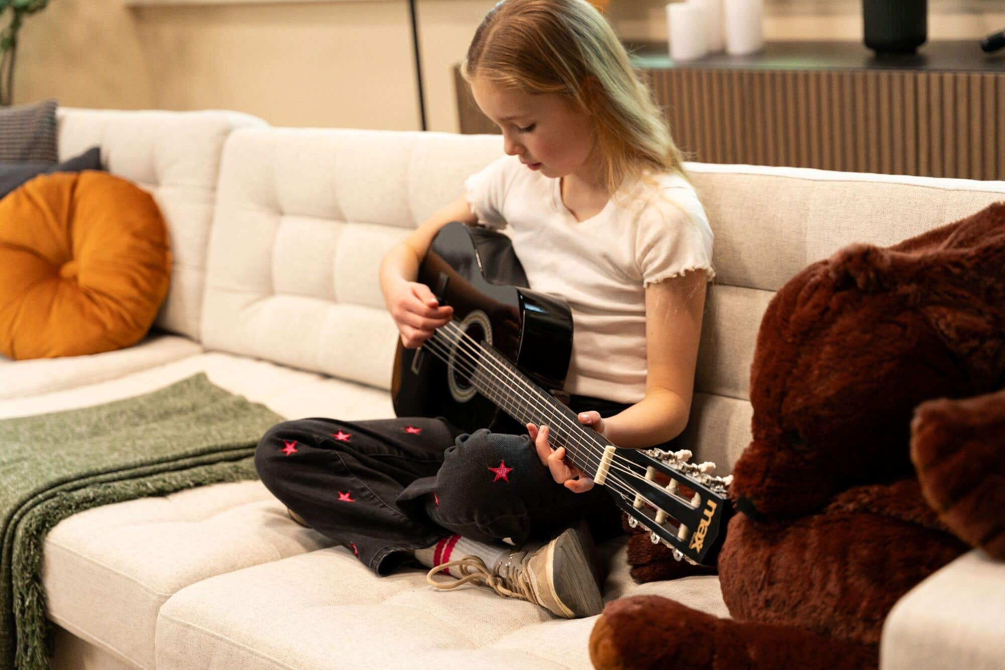 Niña tocando guitarra negra en sofá beige. Usa camisa blanca y pantalón negro con estrellas.
