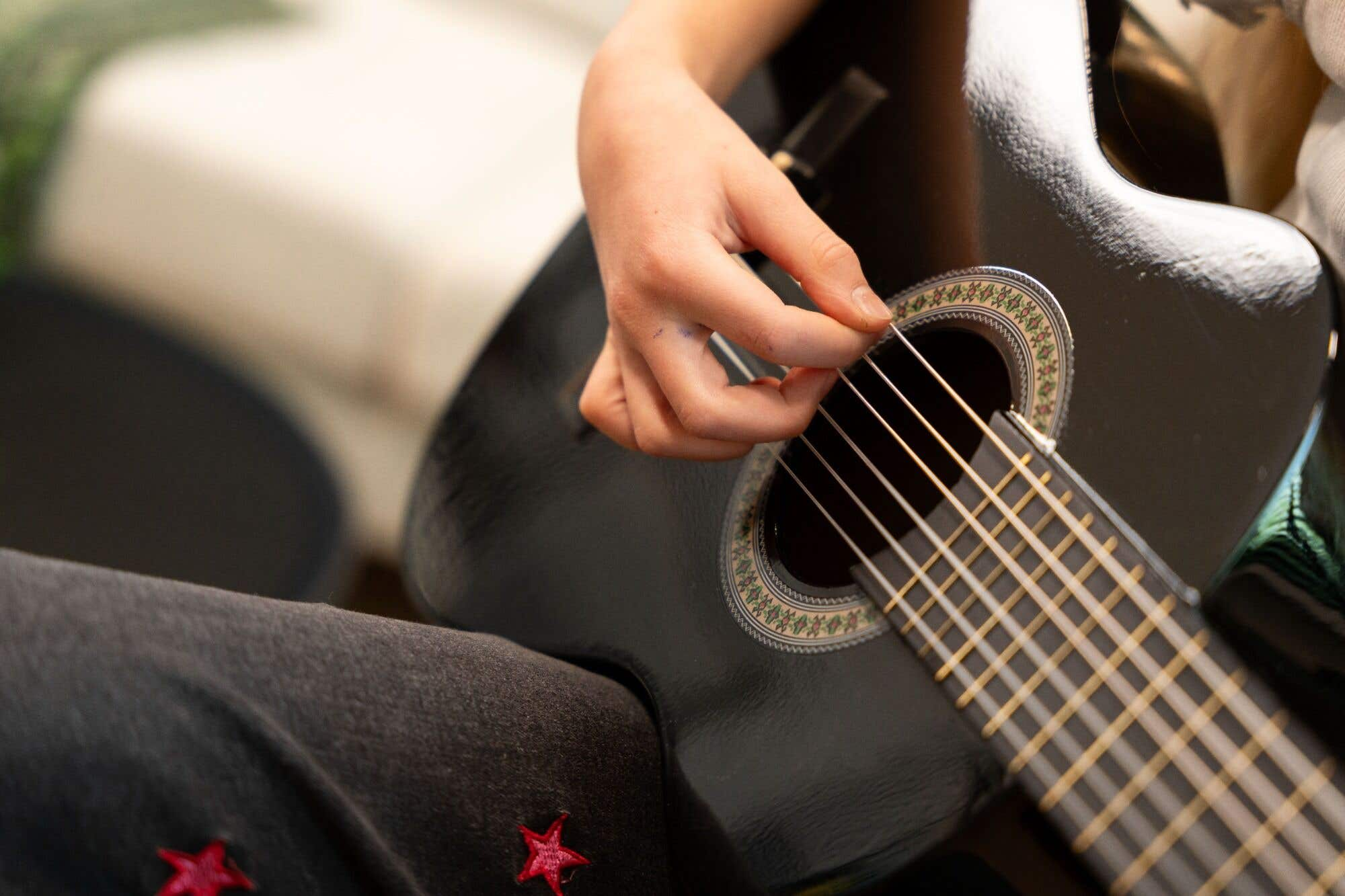 Primer plano de una mano tocando las cuerdas de una guitarra negra. La niña usa un pantalón.
