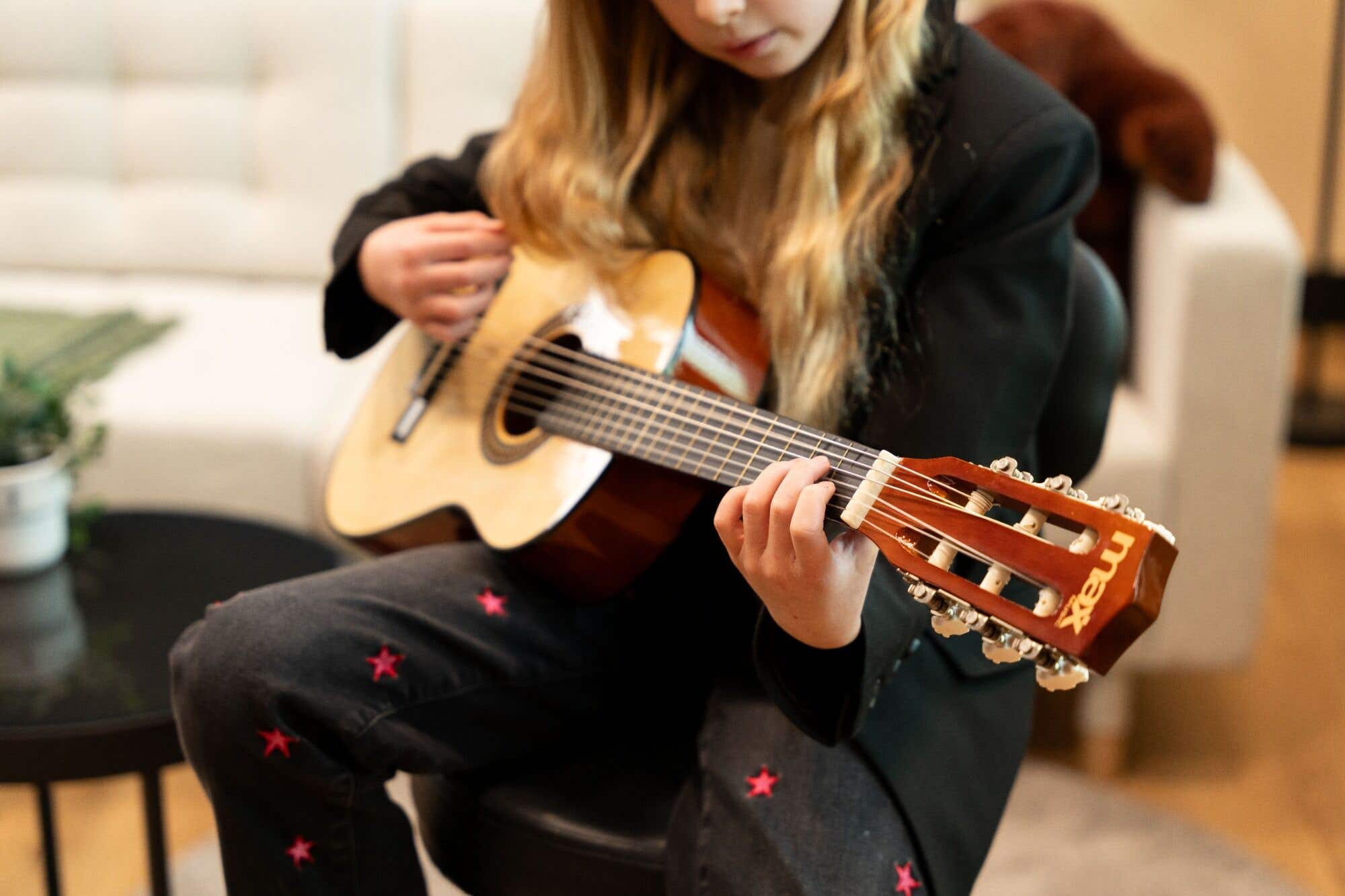 Una niña toca la guitarra, sentada. La guitarra es marrón claro. Lleva una blazer negro y jeans con estrellas rosas.