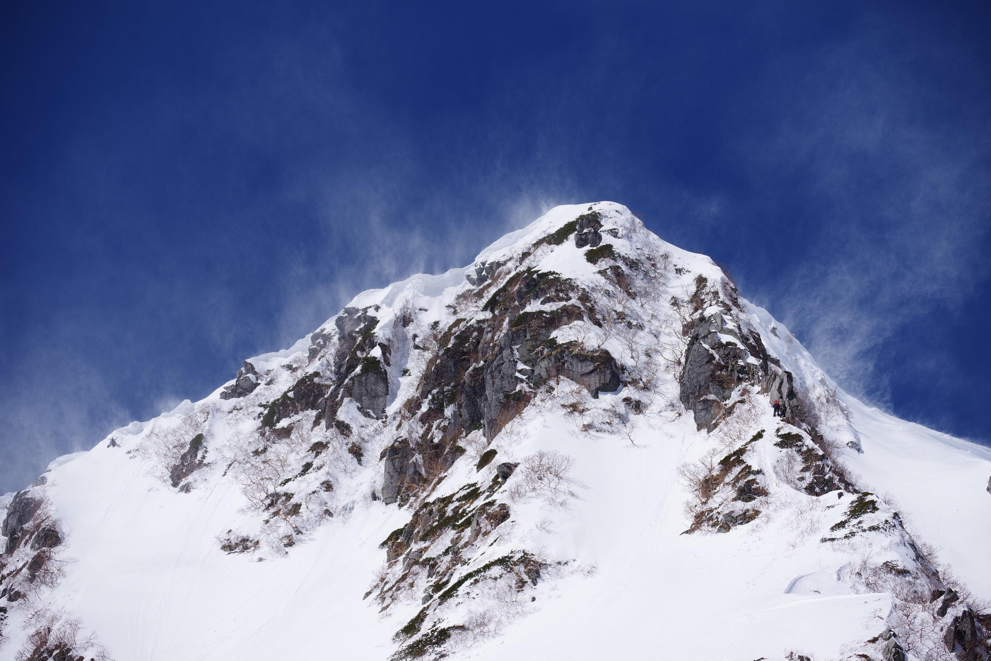 Verschneite Bergspitze vor blauem Himmel. Auf dem Gipfel sind Felsen und grüne Vegetation zu sehen.