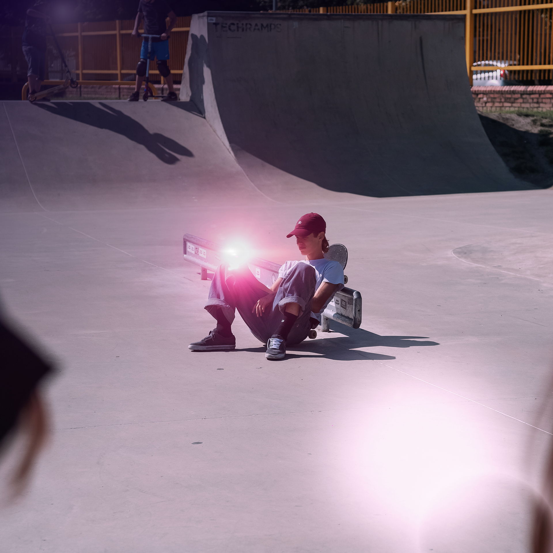 Osoba siedzi na deskorolce w skateparku, z rampami i innymi skaterami w tle.