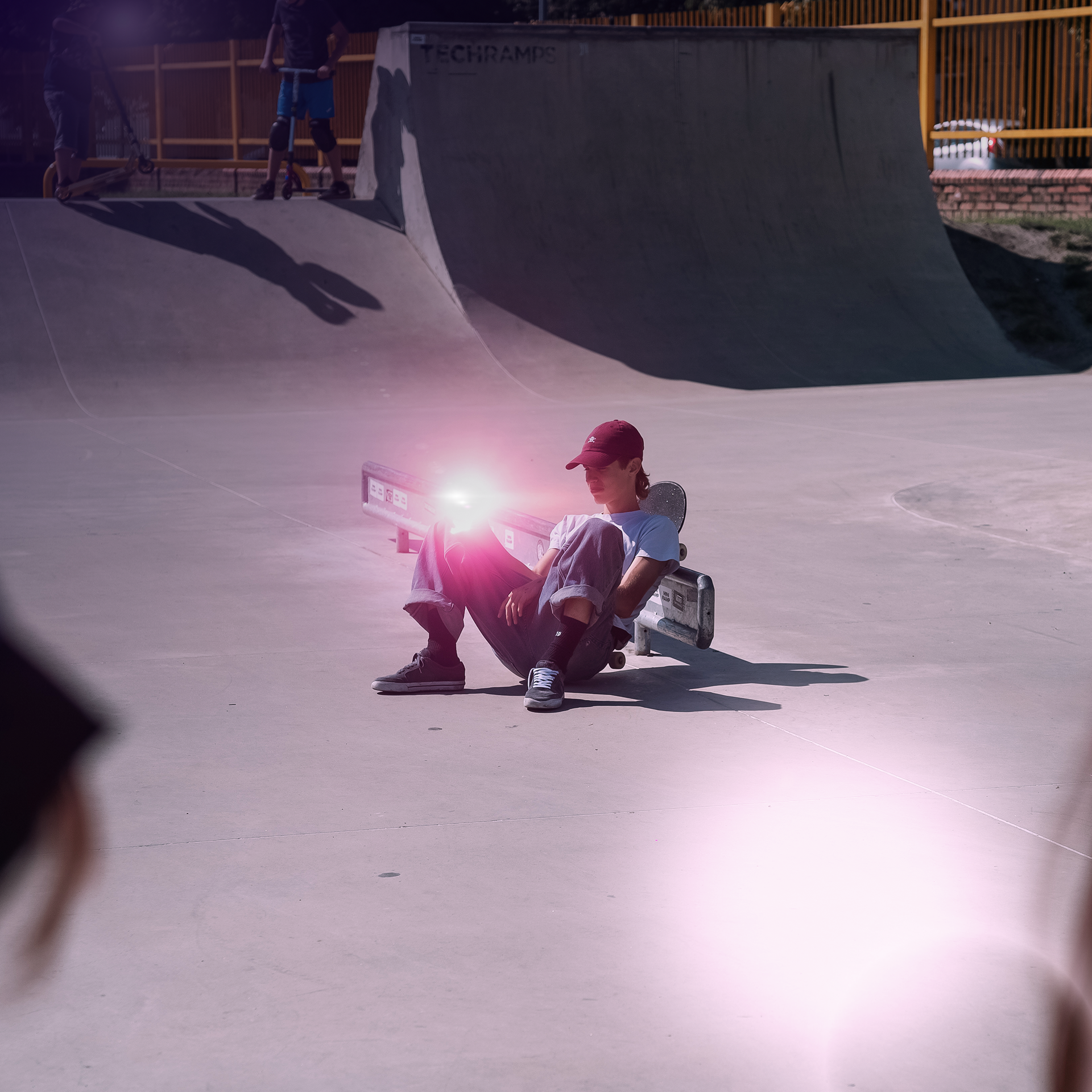 Una persona seduta su una rampa per skateboard, che tiene un telefono. Altre persone sullo sfondo.