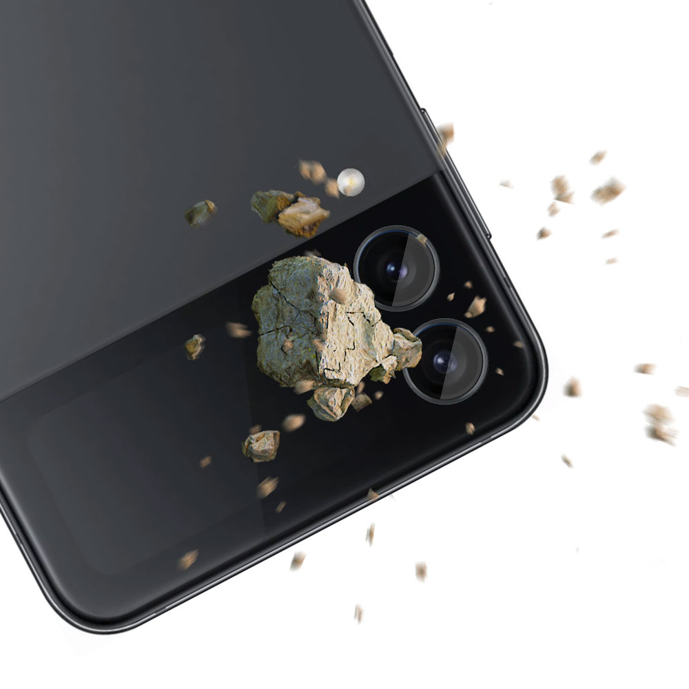 Un telefono nero mostra protezioni per l'obiettivo sotto impatto, con detriti sparsi attorno alla fotocamera a doppia lente su sfondo bianco.