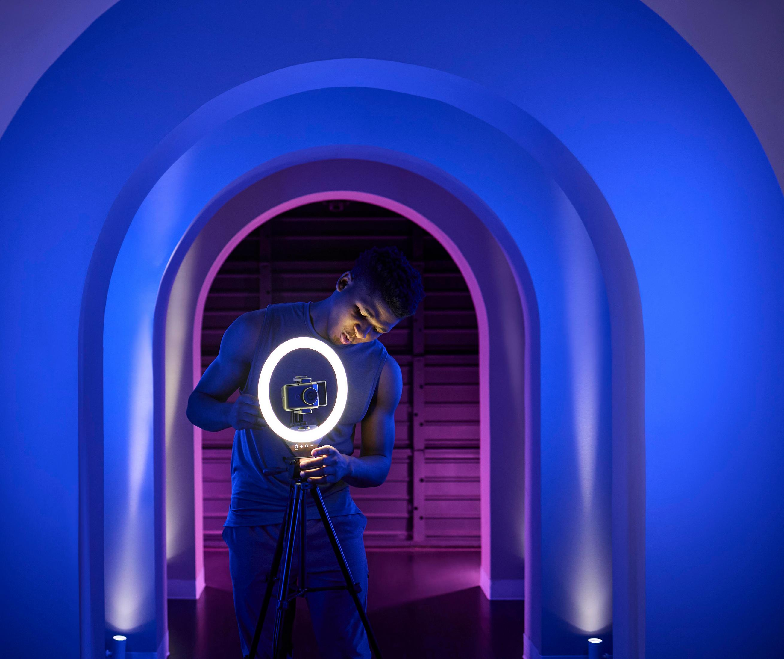 Ein Mann stellt eine Kamera auf einem Stativ in einem blau-violetten Raum mit Ringlichtern auf.