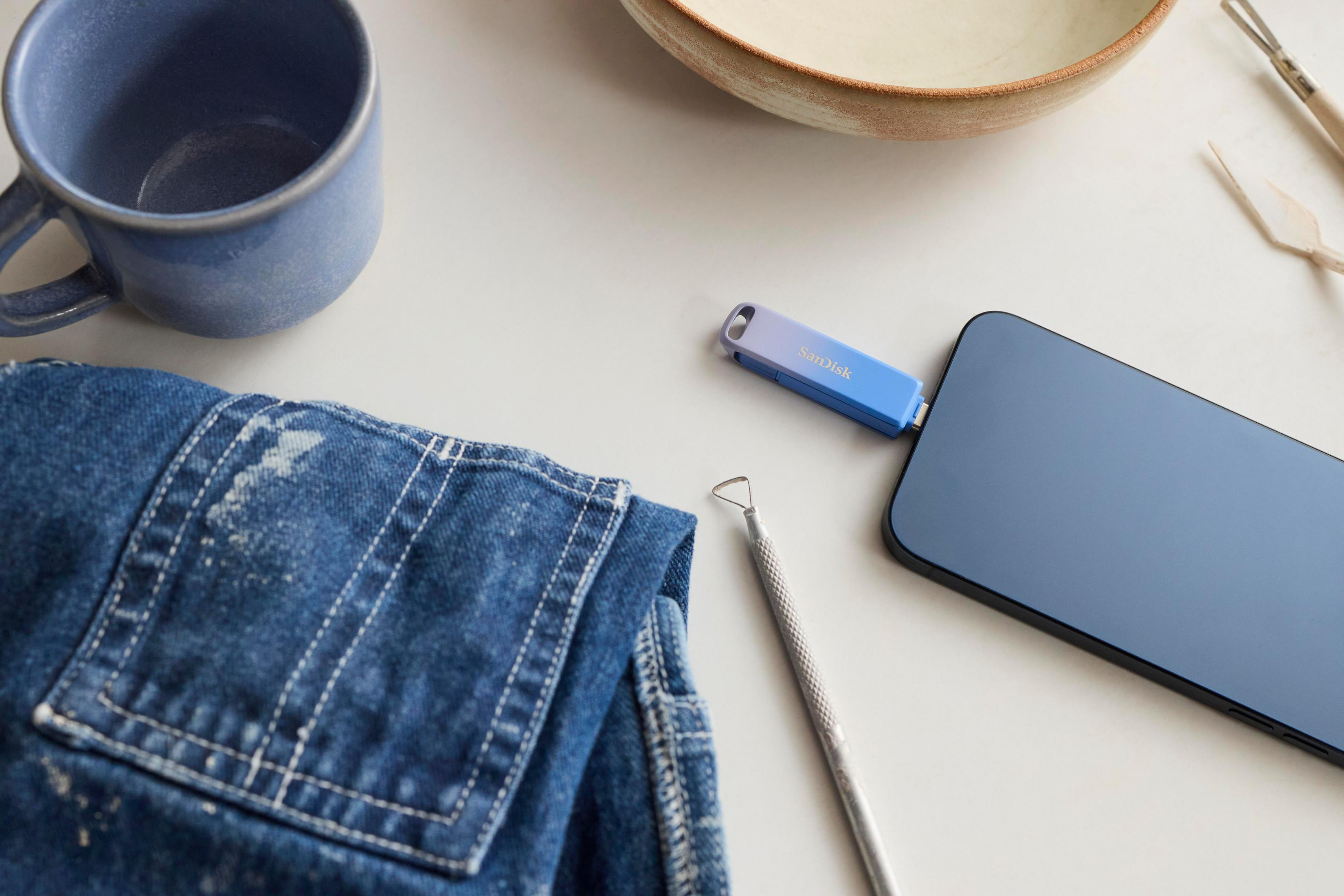 Ten obraz przedstawia pendrive SanDisk, telefon, kubek i kilka narzędzi na białej powierzchni z dżinsem.