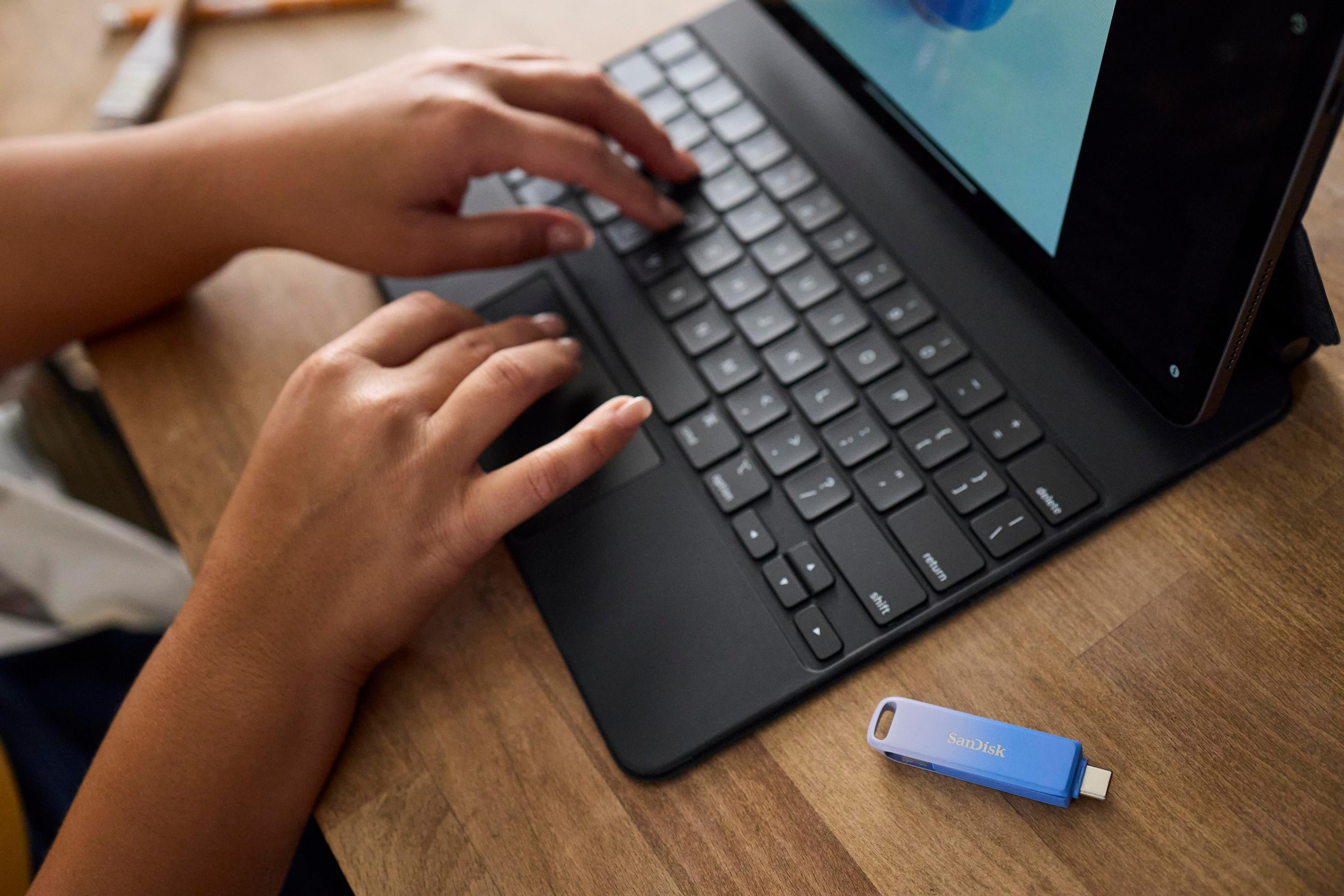 Obraz przedstawia pendrive SanDisk obok czarnej klawiatury i tabletu na drewnianym stole.