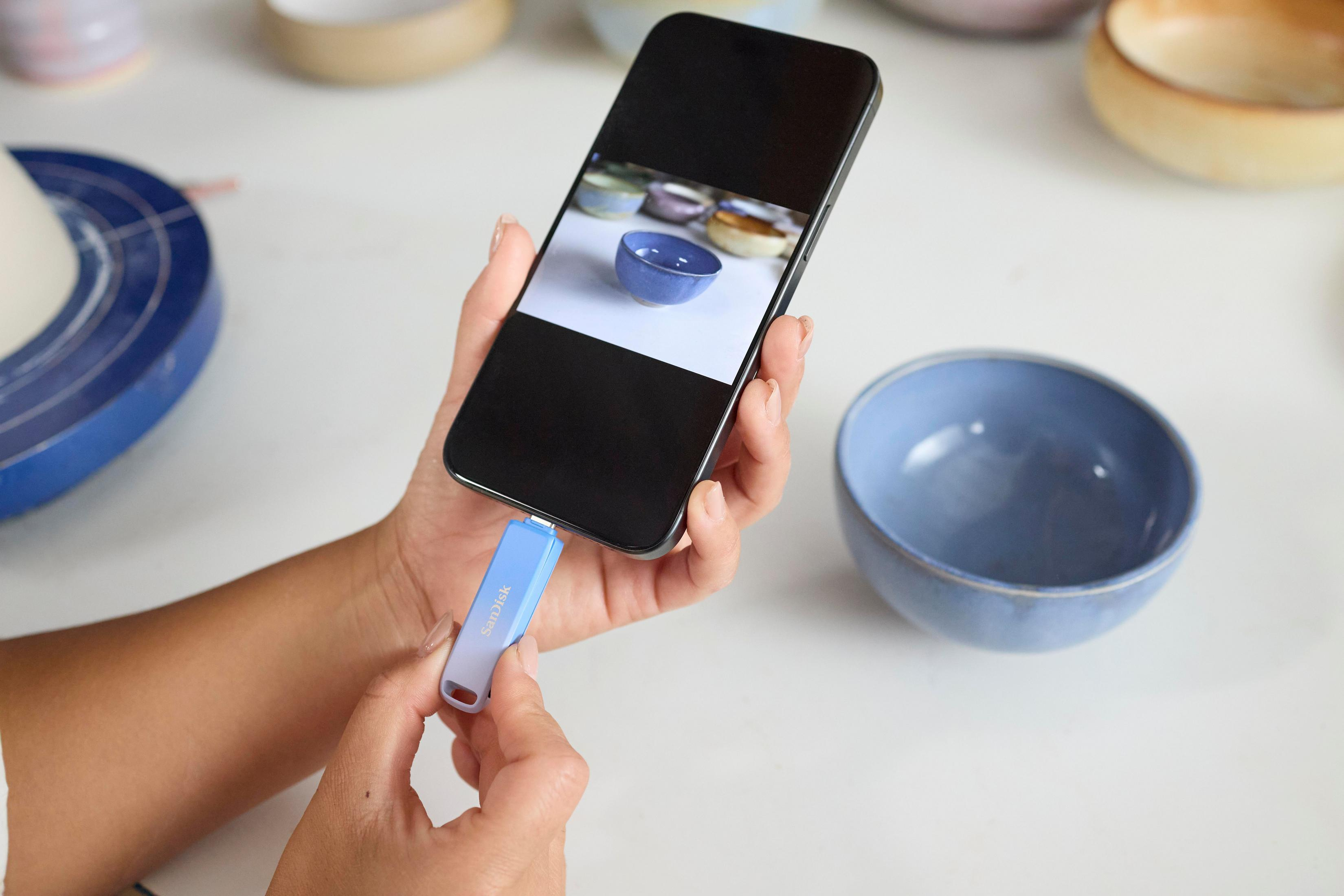 Obraz przedstawia rękę podłączającą pendrive SanDisk do telefonu, obok misek i biurka.